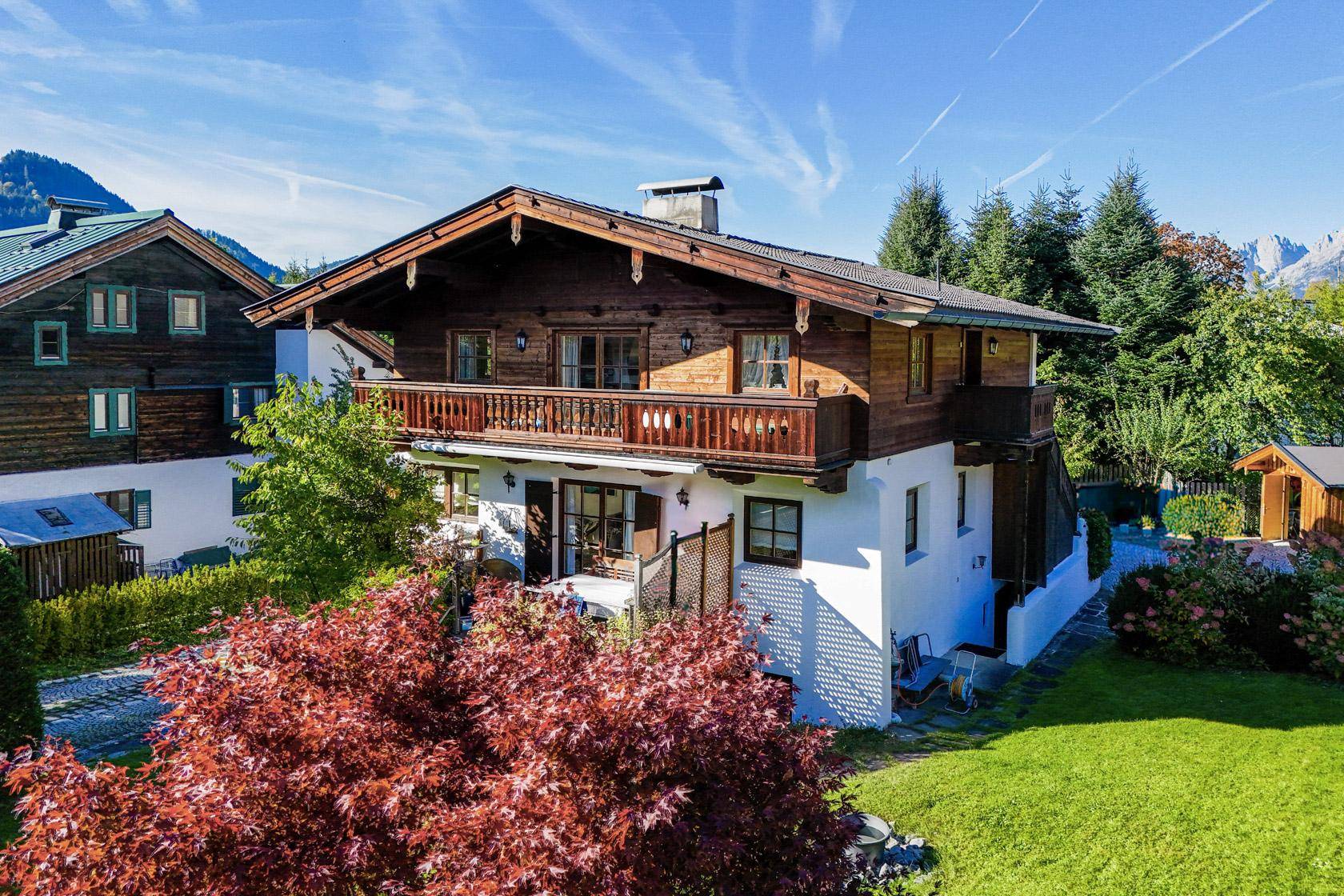 Charmantes Alpenhaus mit Holzbalkonen, gepflegtem Garten und einem leuchtend roten Baum im Vordergrund.