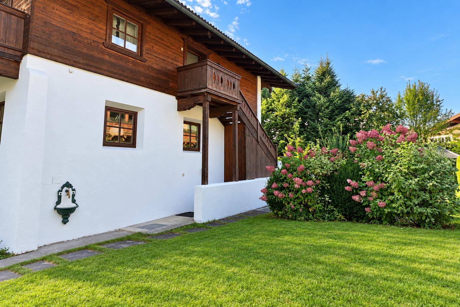Seitenansicht des Hauses mit weißer Fassade, Holztreppe zum Balkon und blühenden Büschen im Garten.