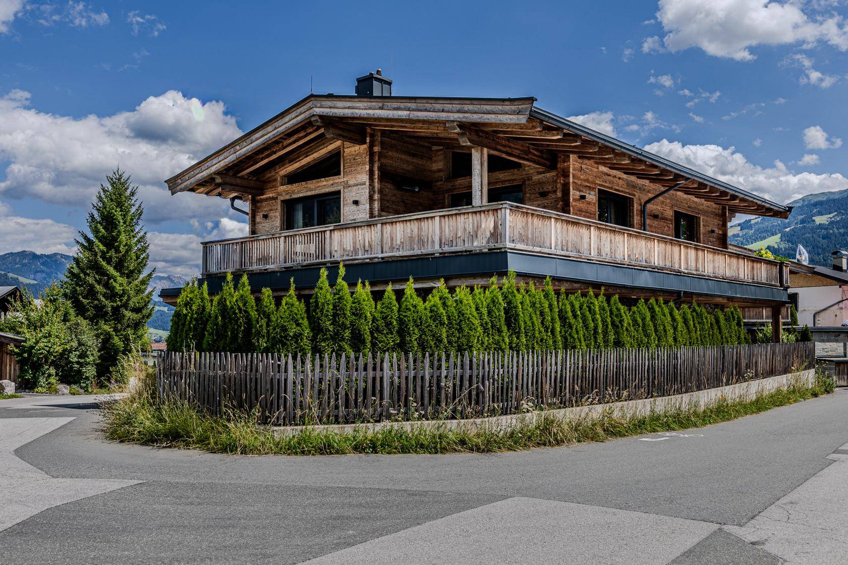 Charmantes Holzchalet mit großem Balkon, umgeben von einer gepflegten Hecke und Alpenlandschaft.