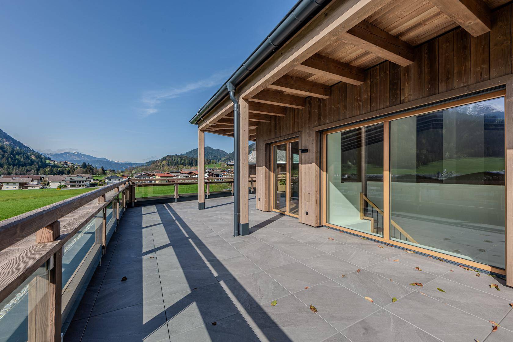 Lange geflieste Terrasse mit Holzfassade und weitem Blick über das Dorf und die Landschaft.