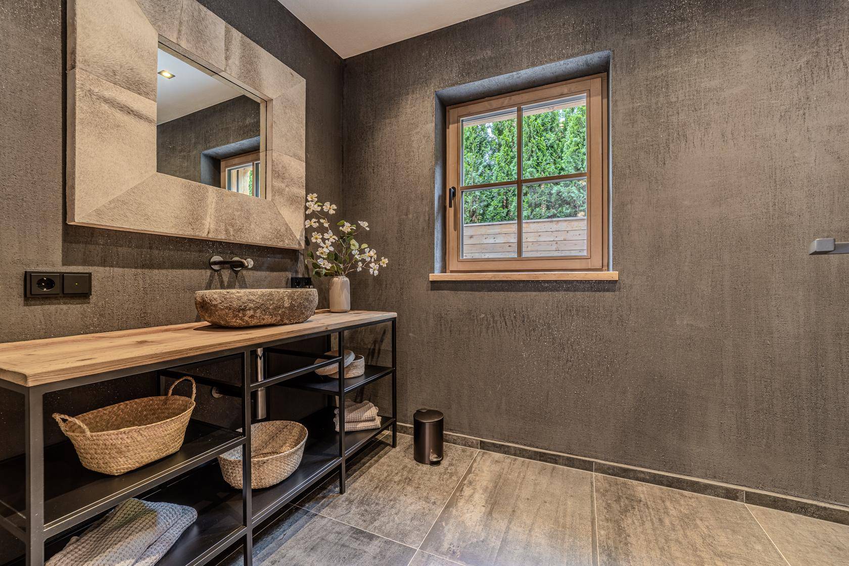 Modernes Badezimmer mit dunklen Fliesen, Holzwaschtisch und Fenster mit Blick ins Grüne.