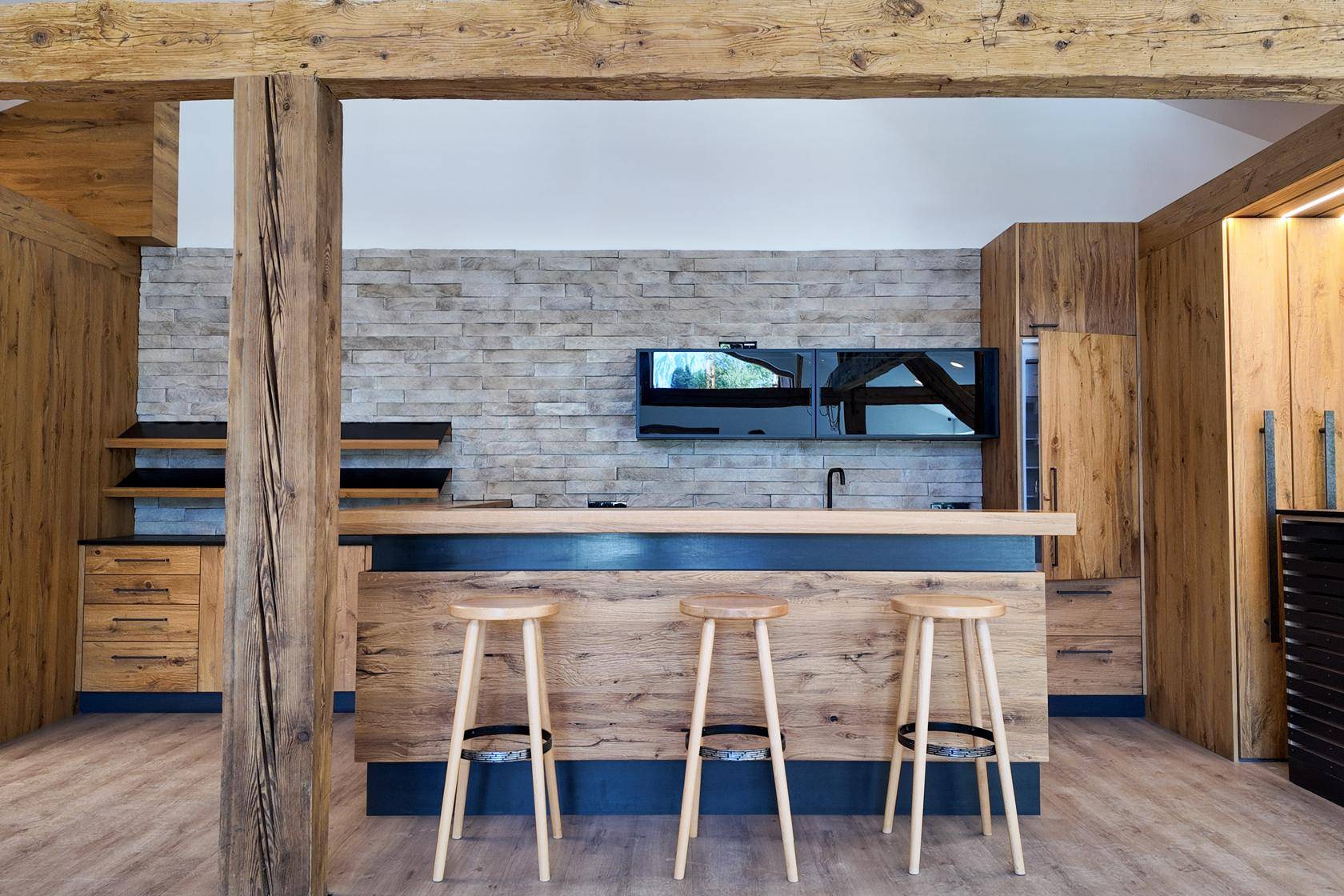Detailansicht einer modernen Küche mit Holzfronten, Steinwand und einer Bar mit drei Hockern.