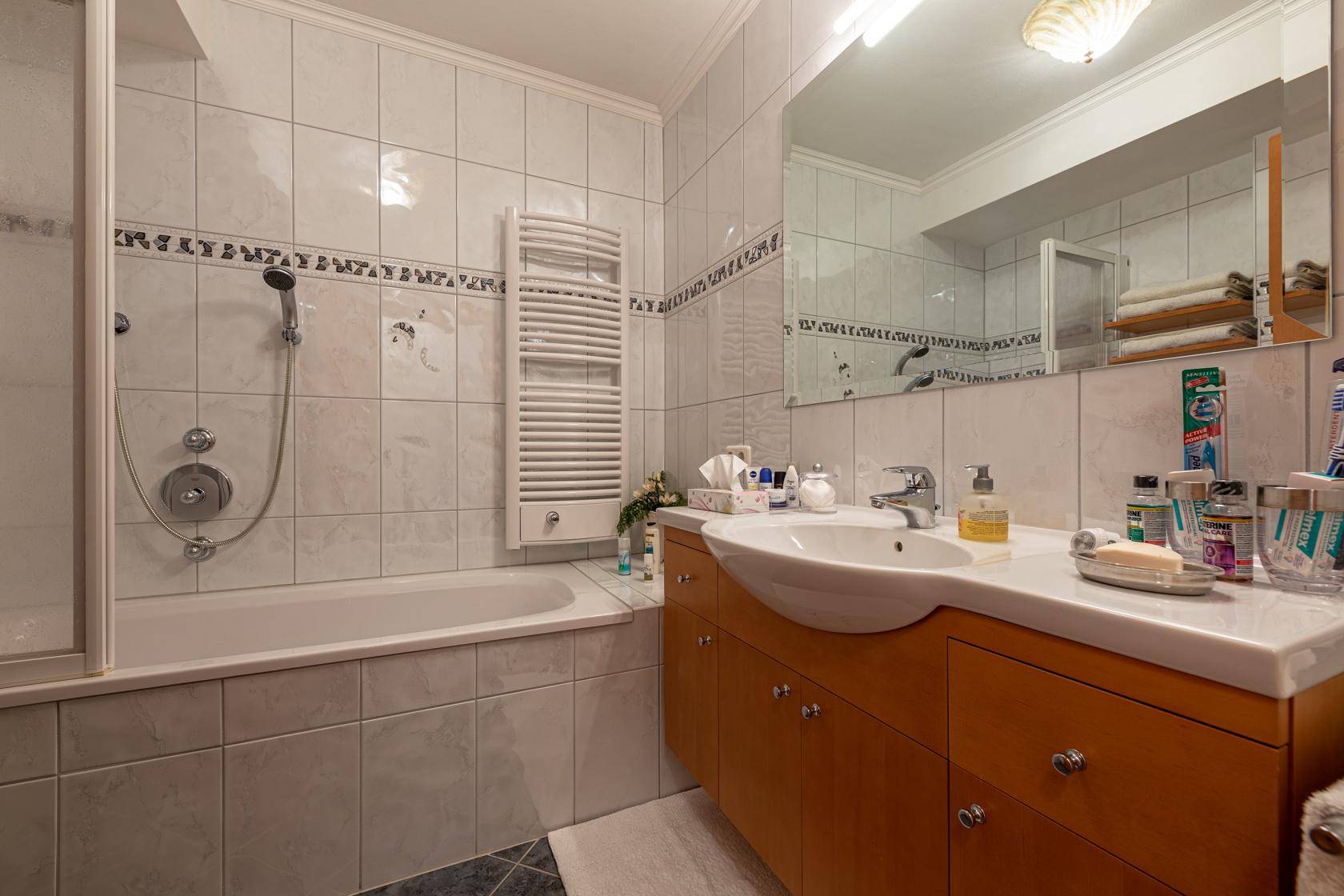 Helles Badezimmer mit Badewanne, Duschfunktion, Waschbecken und beheiztem Handtuchhalter.