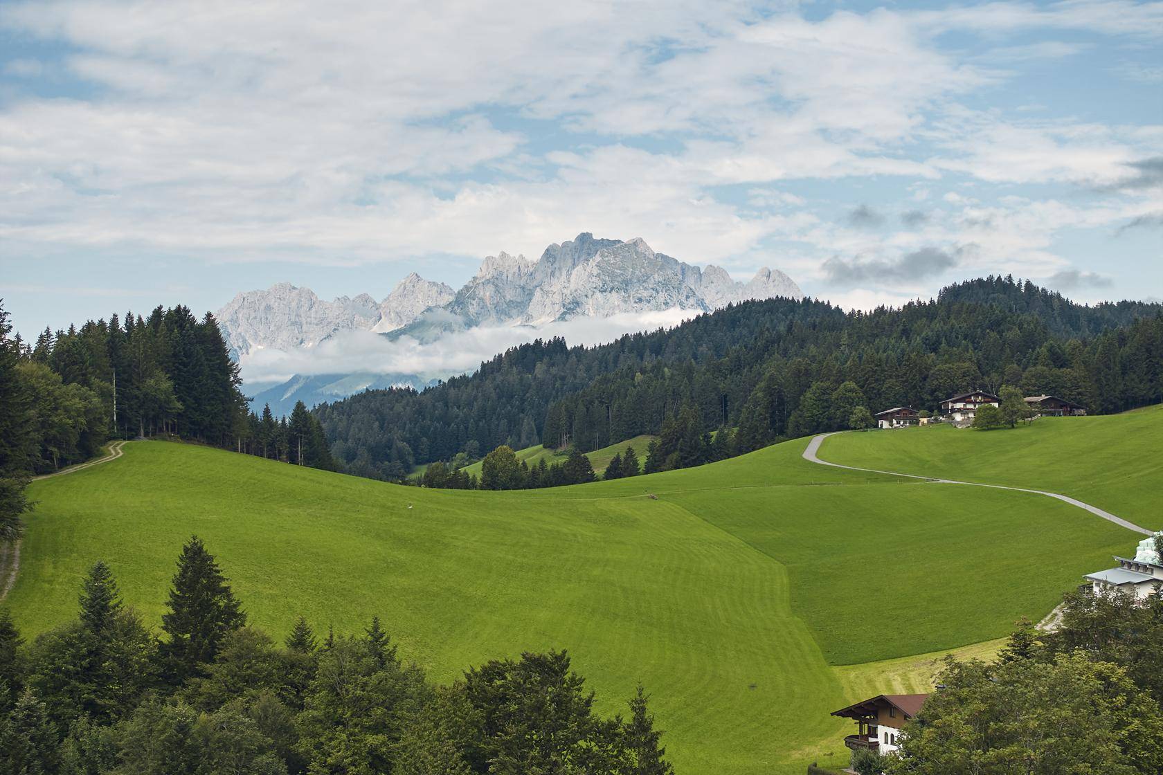 Grüne Hügellandschaft mit vereinzelten Häusern und majestätischen Bergen am Horizont.
