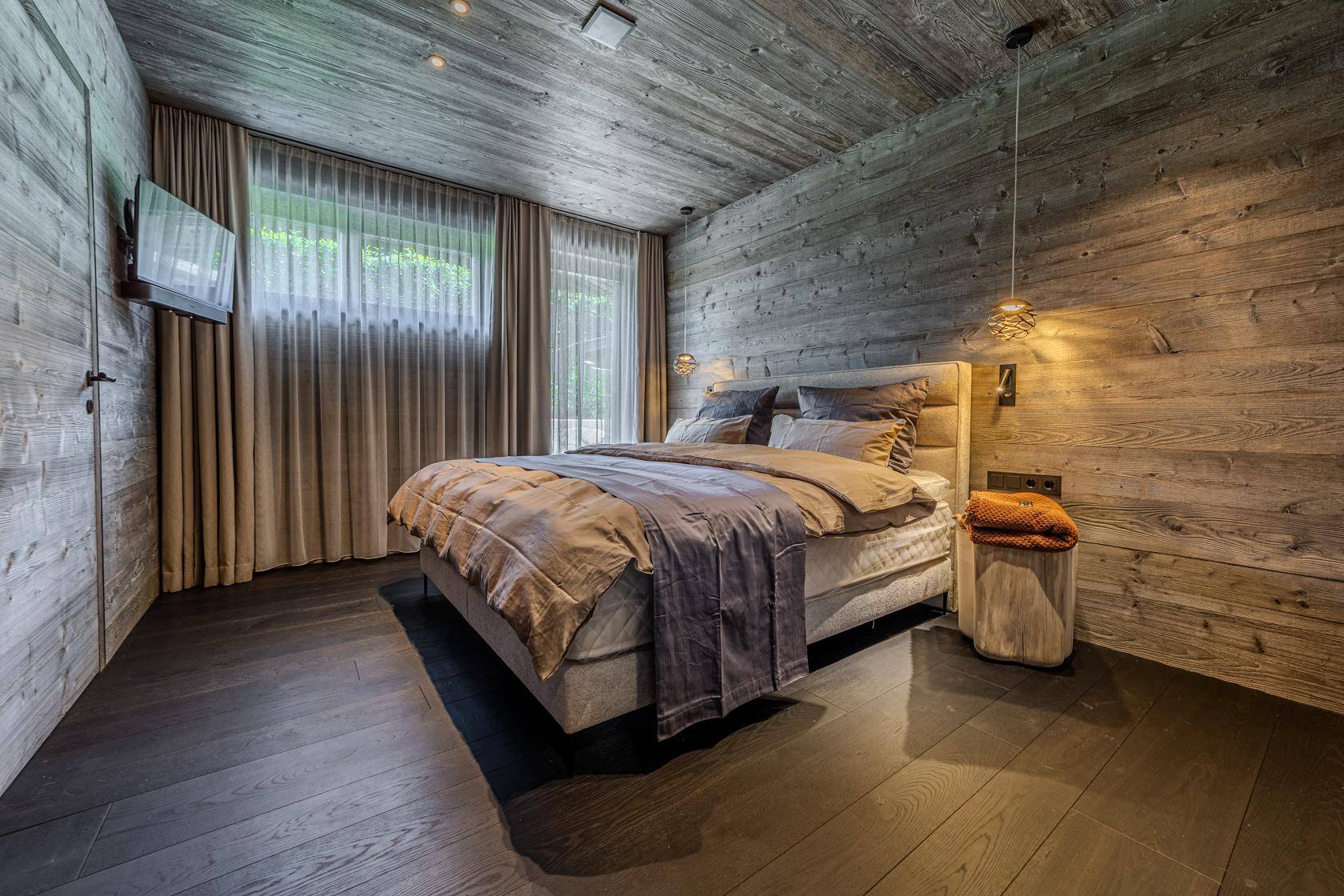 Gemütliches Schlafzimmer mit Holzwänden, Holzbalkendecke und einem Fenster mit blickdichten Vorhängen.