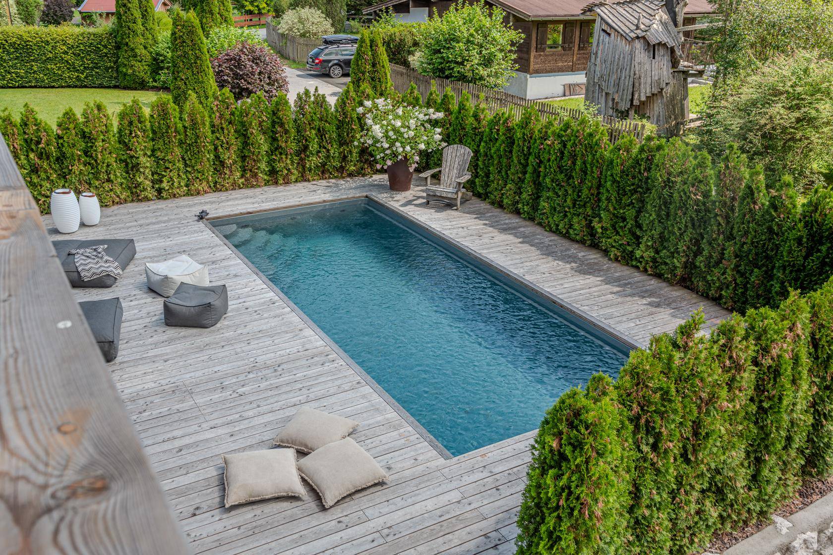 Außenbereich mit rechteckigem Pool und Holzdeck, umgeben von einer dichten Hecke und Lounge-Kissen.