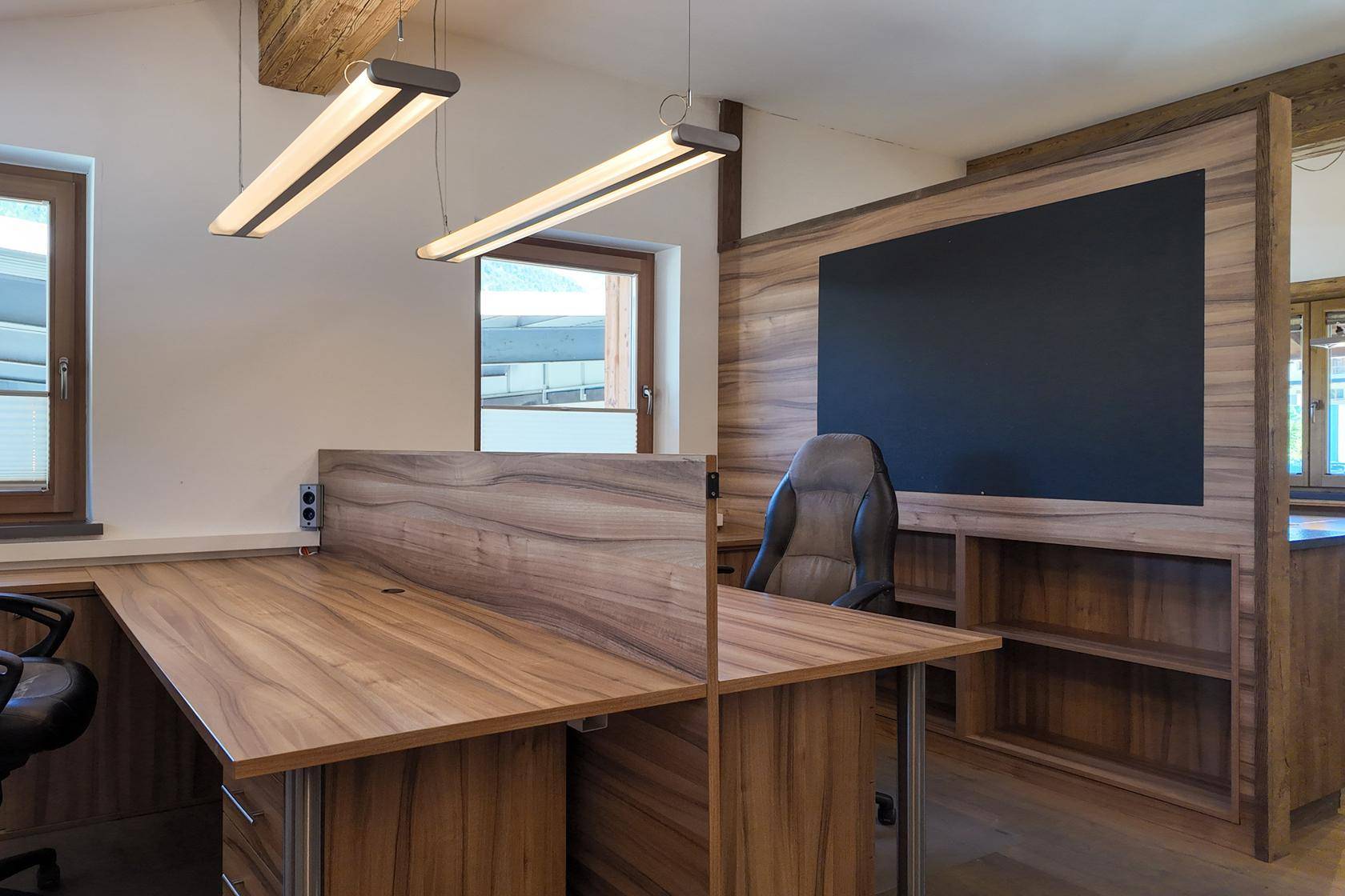 Büroarbeitsplatz mit Holzschreibtisch, Bürostuhl und integrierten Regalen, beleuchtet durch Deckenleuchten.