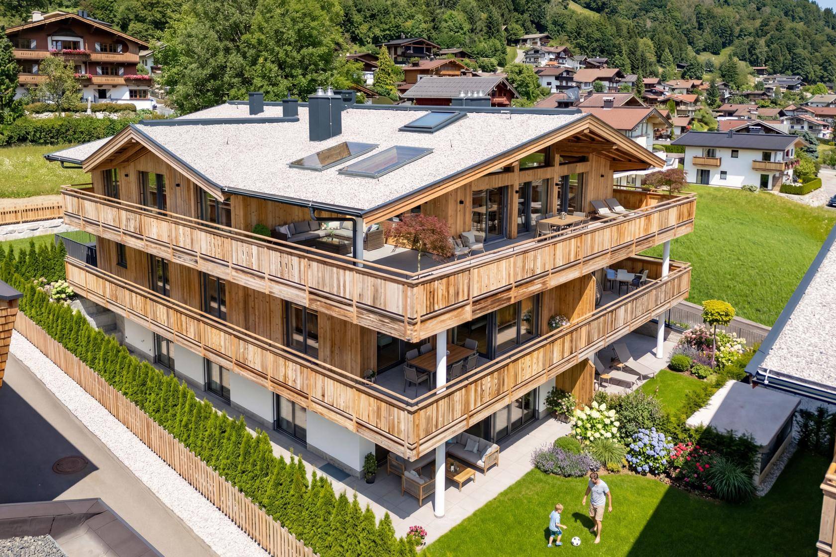 Modernes Holzchalet mit mehreren Balkonen und Terrassen, umgeben von grüner Berglandschaft und Rasenflächen.