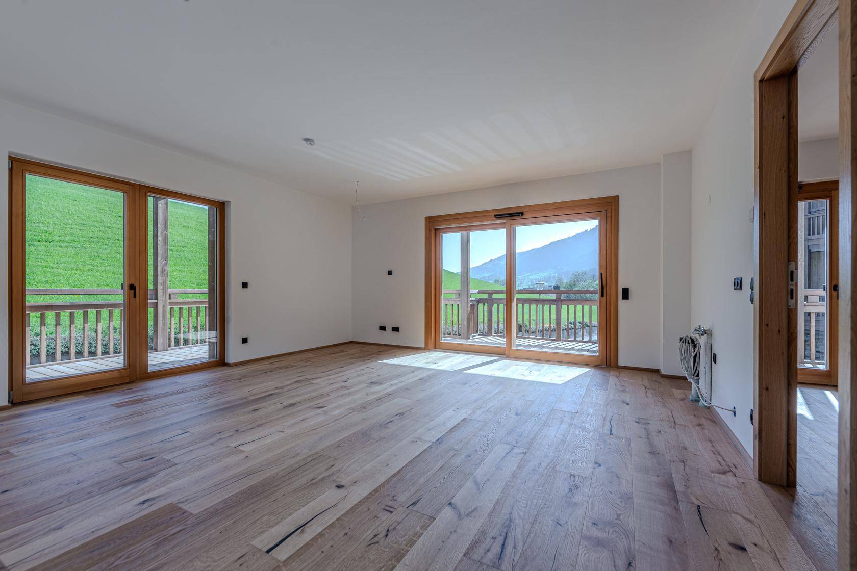 Helles Wohnzimmer mit Holzboden und großen Fenstern, die einen weiten Blick ins Grüne und auf die Berge bieten.