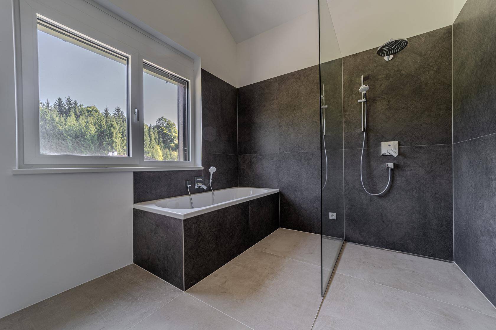 Modernes Badezimmer mit dunklen Fliesen, Badewanne, begehbarer Dusche und Fenster mit Waldblick.