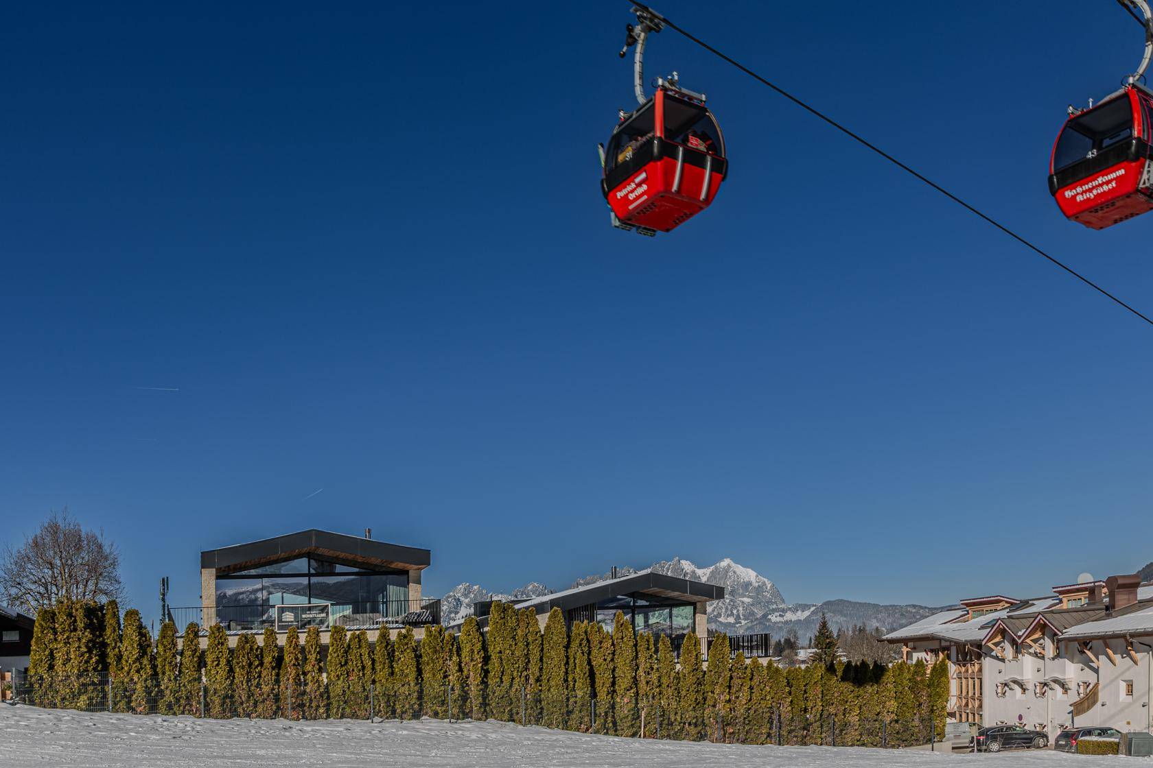Moderne Architektur mit großen Glasflächen und einer Hecke im Vordergrund, umgeben von Schnee und Bergen.