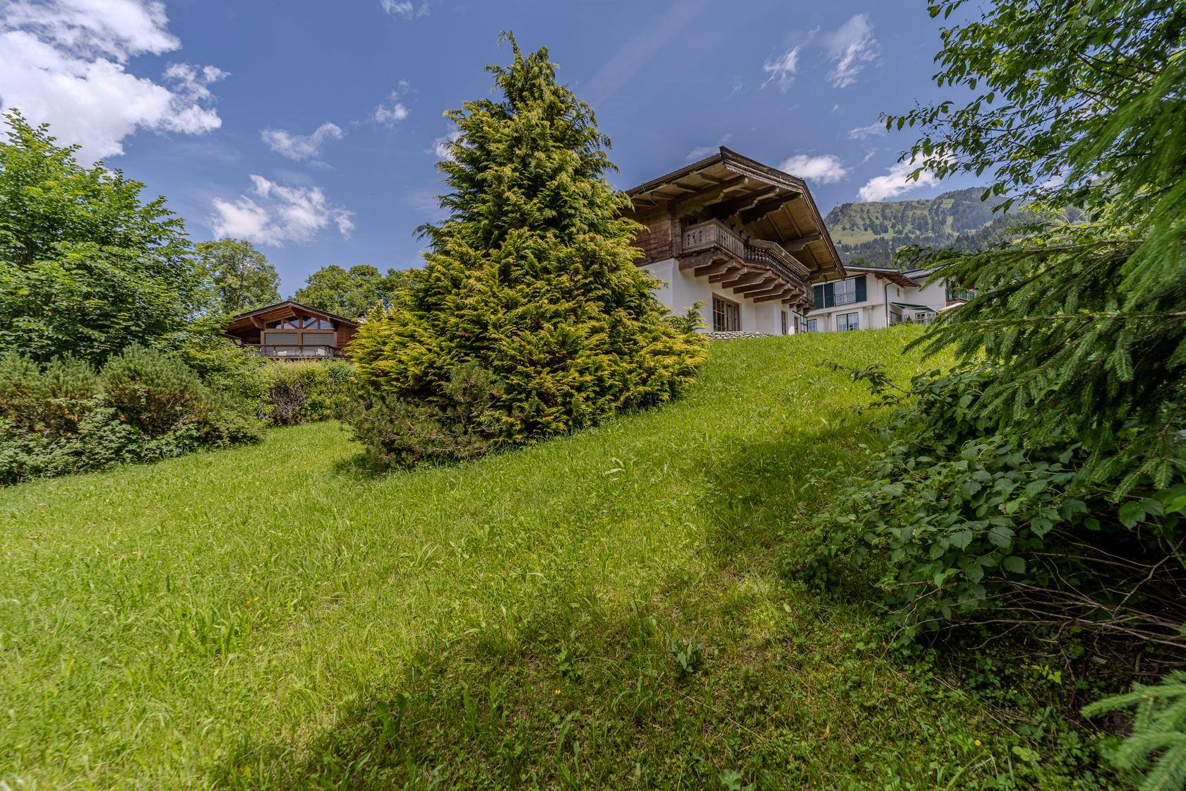 Weitläufige Grünfläche mit Bäumen und Blick auf das charmante Alpenhaus im Hintergrund.