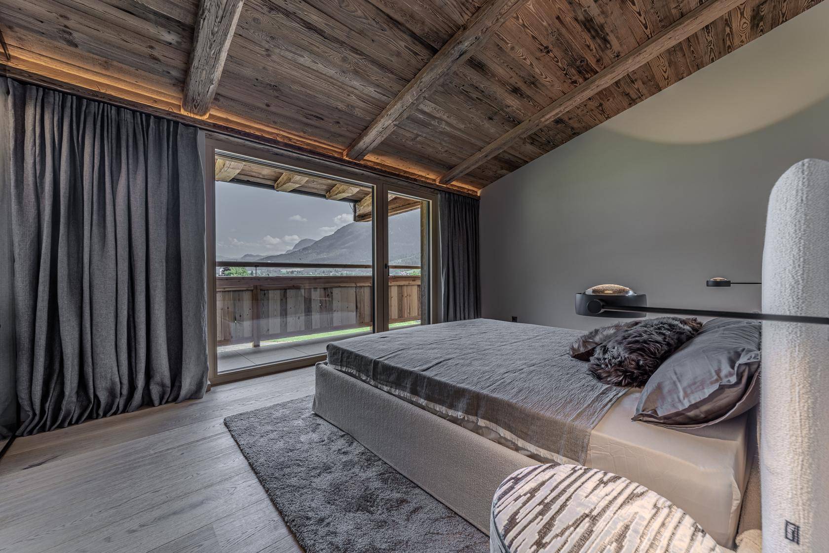Gemütliches Schlafzimmer mit Holzbalkendecke, großen Fenstern und weitem Bergblick.
