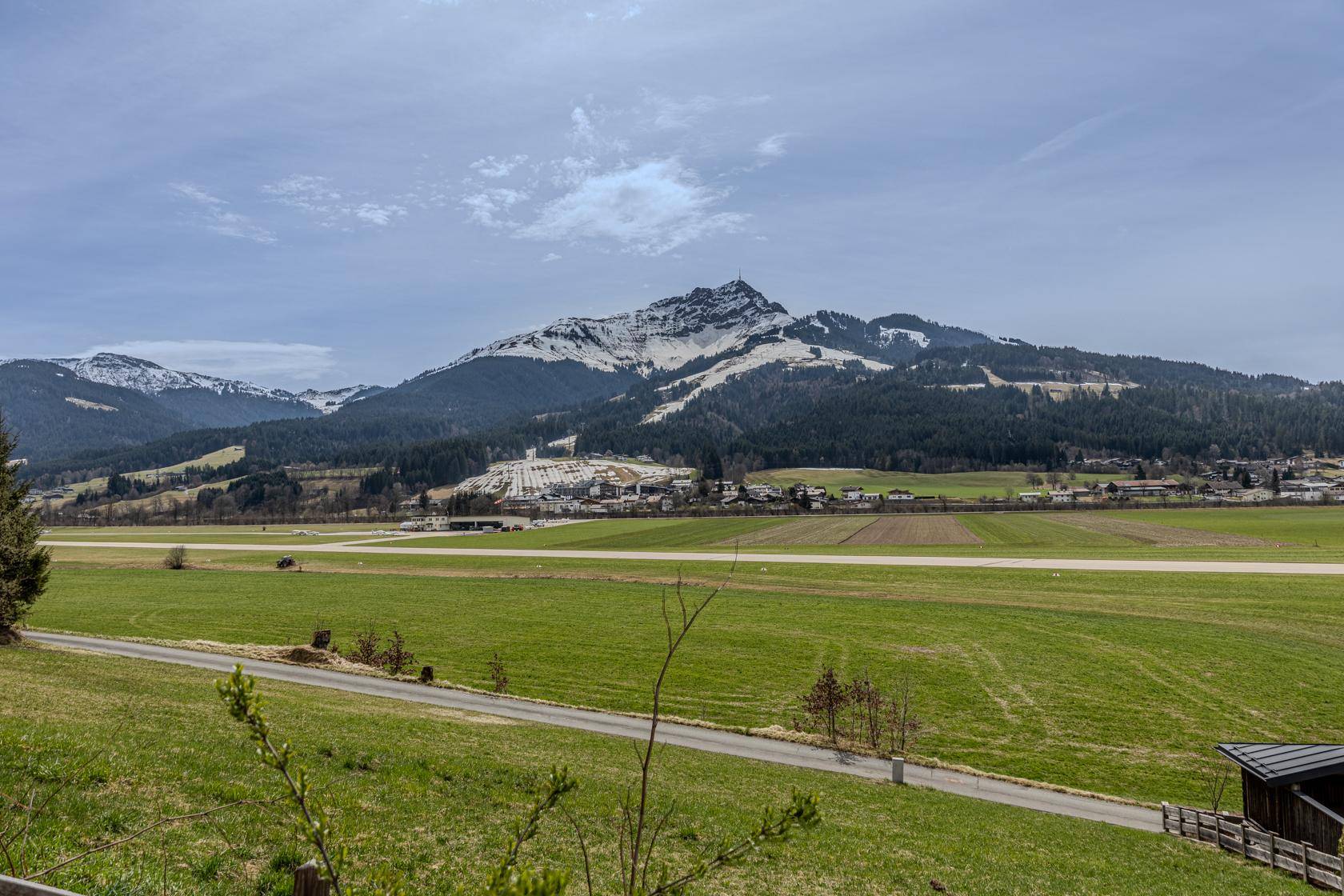 Breiter Blick über grüne Wiesen und Felder mit einem schneebedeckten Bergmassiv im Hintergrund unter hellem Himmel.