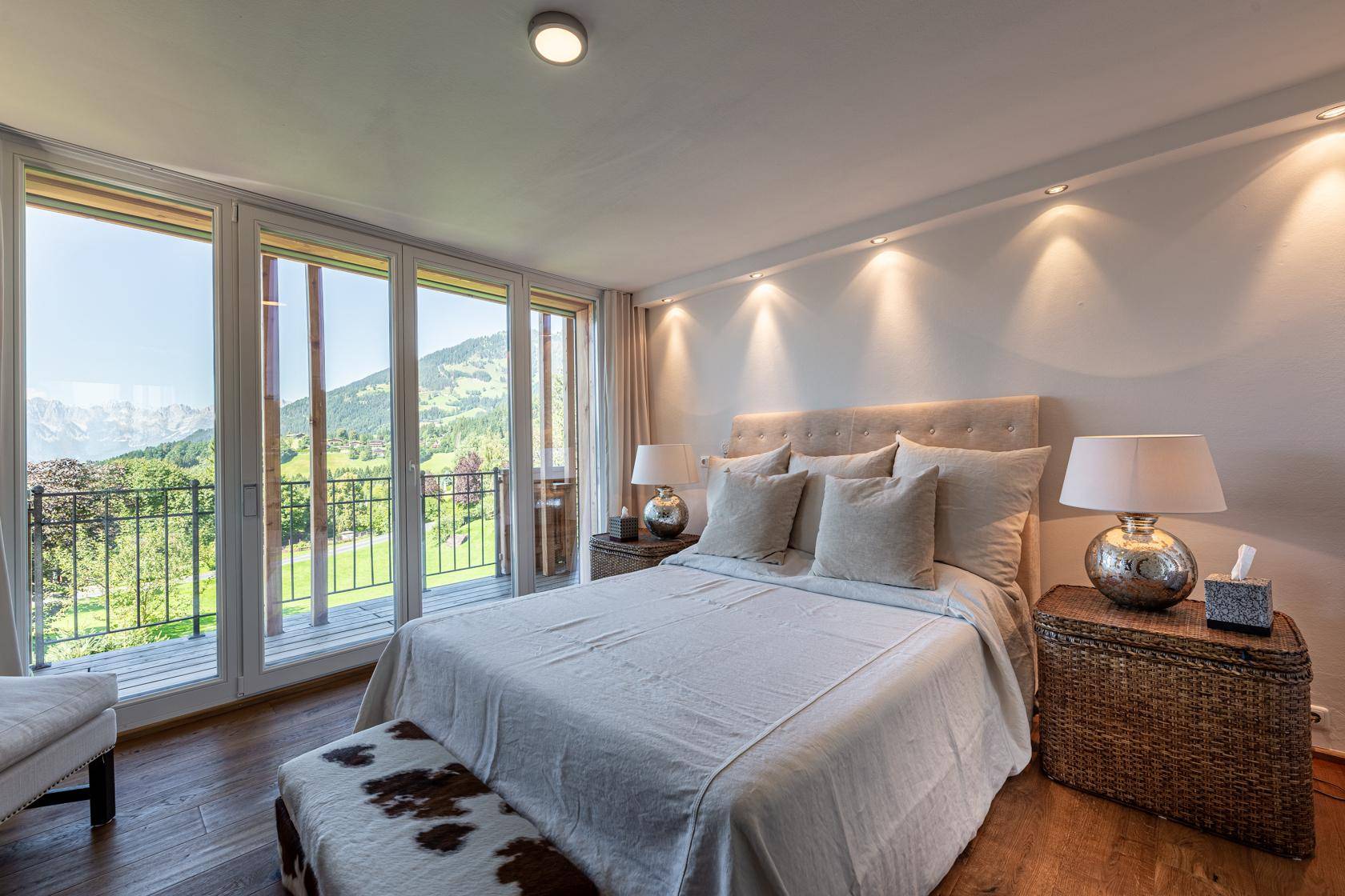 Helles Schlafzimmer mit Holzboden, großen Fensterfronten und Balkonzugang mit Bergblick.