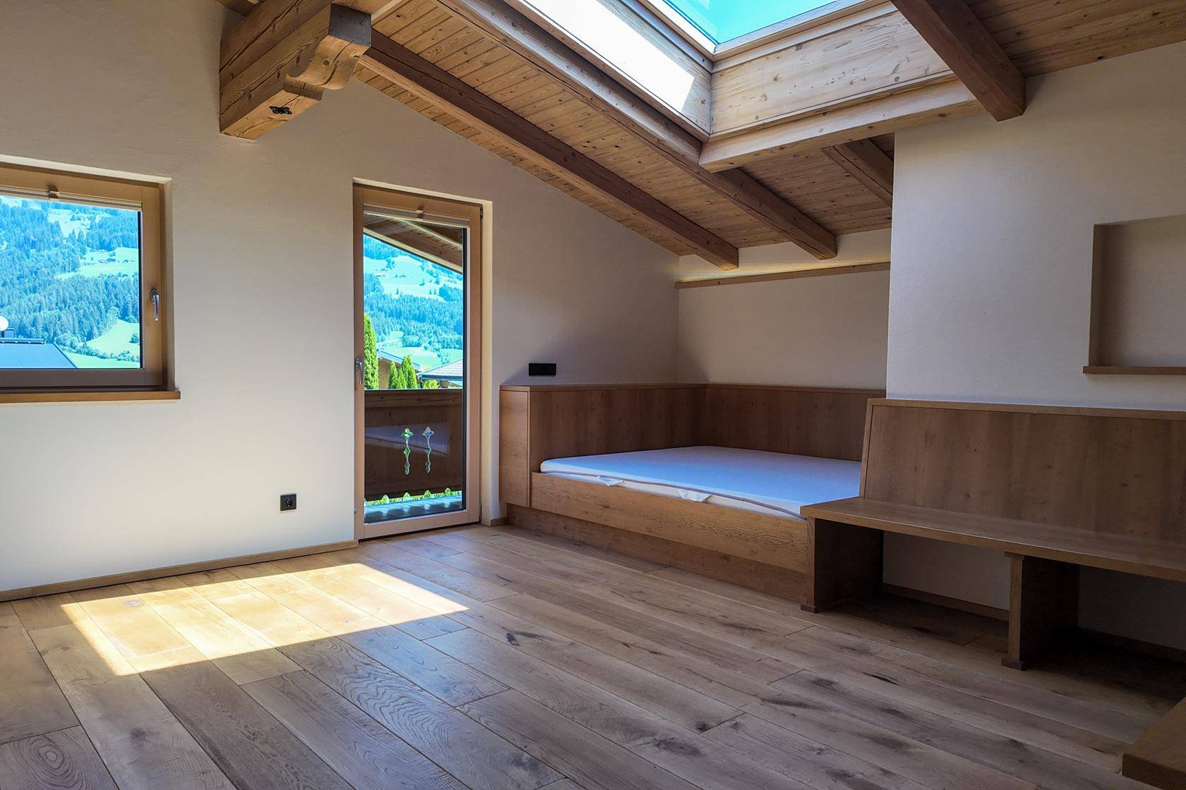 Heller Wohnbereich mit Holzbalkendecke, Einbaubänken und Bergblick durch die Balkontür.