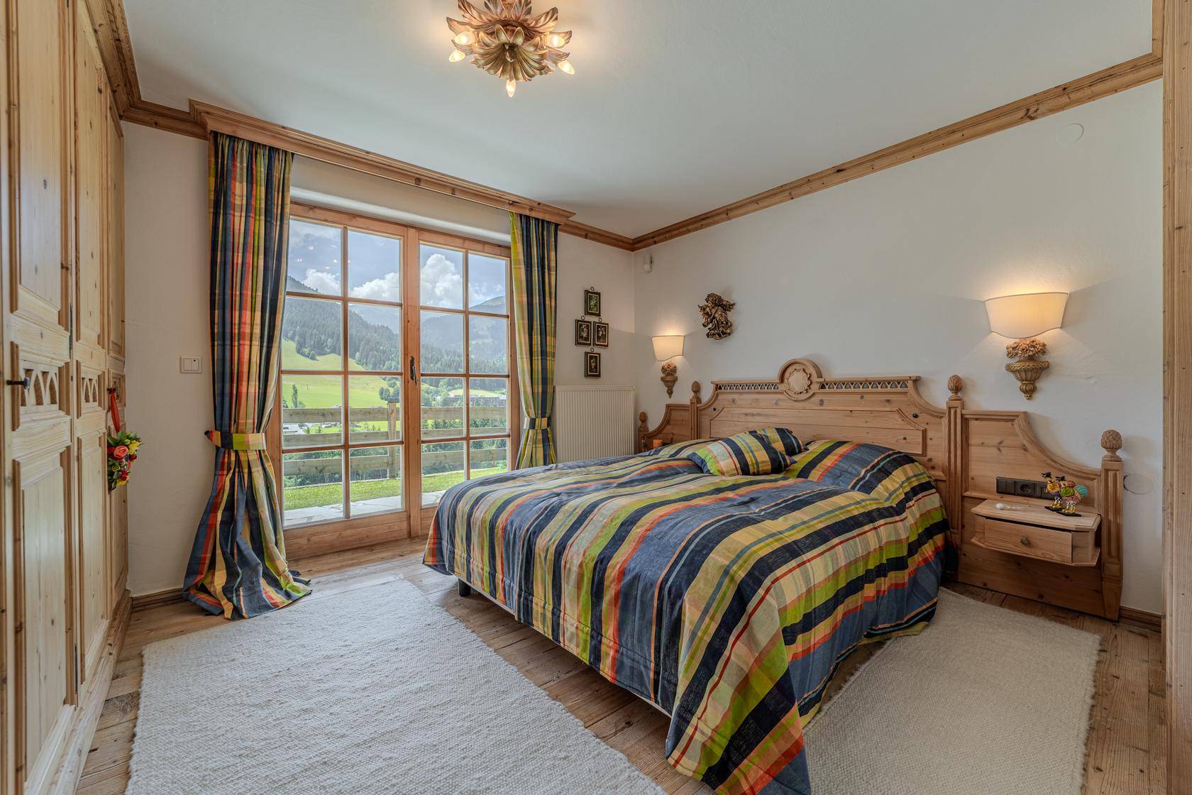 Helles Schlafzimmer mit karierter Bettwäsche, Holzmöbeln und Zugang zu einem Balkon mit Bergblick.