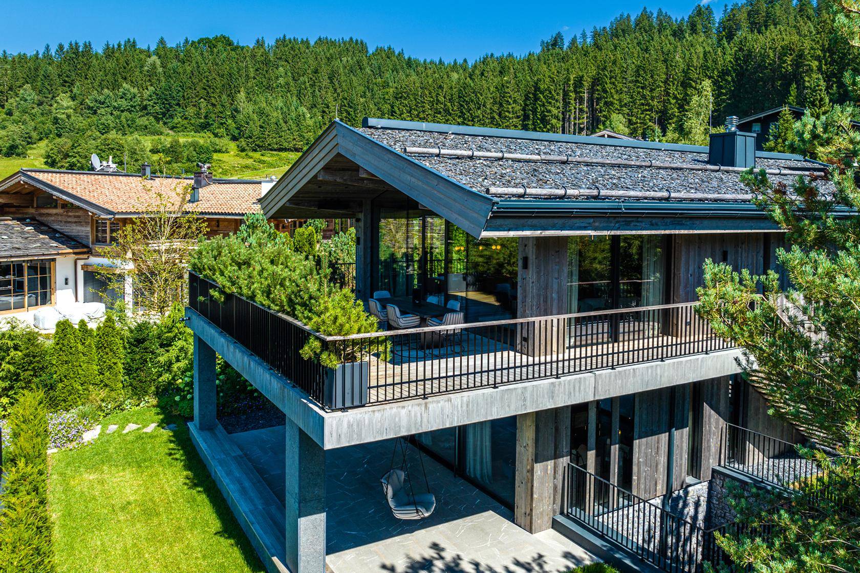 Modernes Chalet mit großem Balkon, umgeben von üppiger grüner Landschaft und Wald.