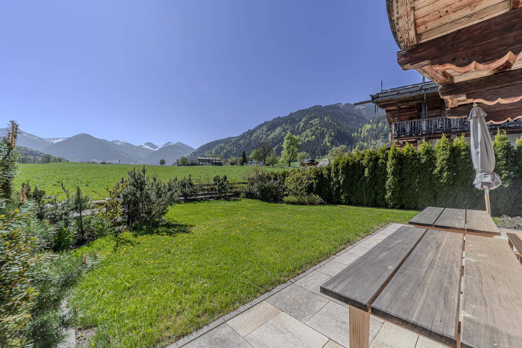 Sonnige Terrasse mit Holztisch und Bänken, angrenzendem Garten und weitem Bergblick.
