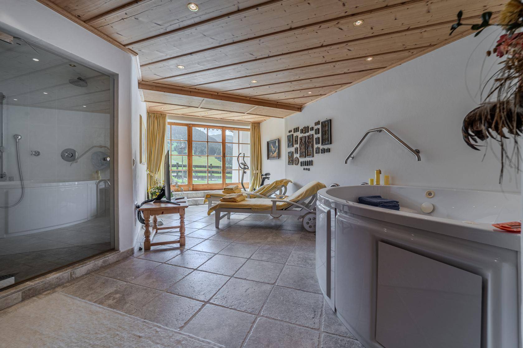 Wellnessbereich mit Whirlpool, Dusche, Liegen und Blick auf die grüne Landschaft.