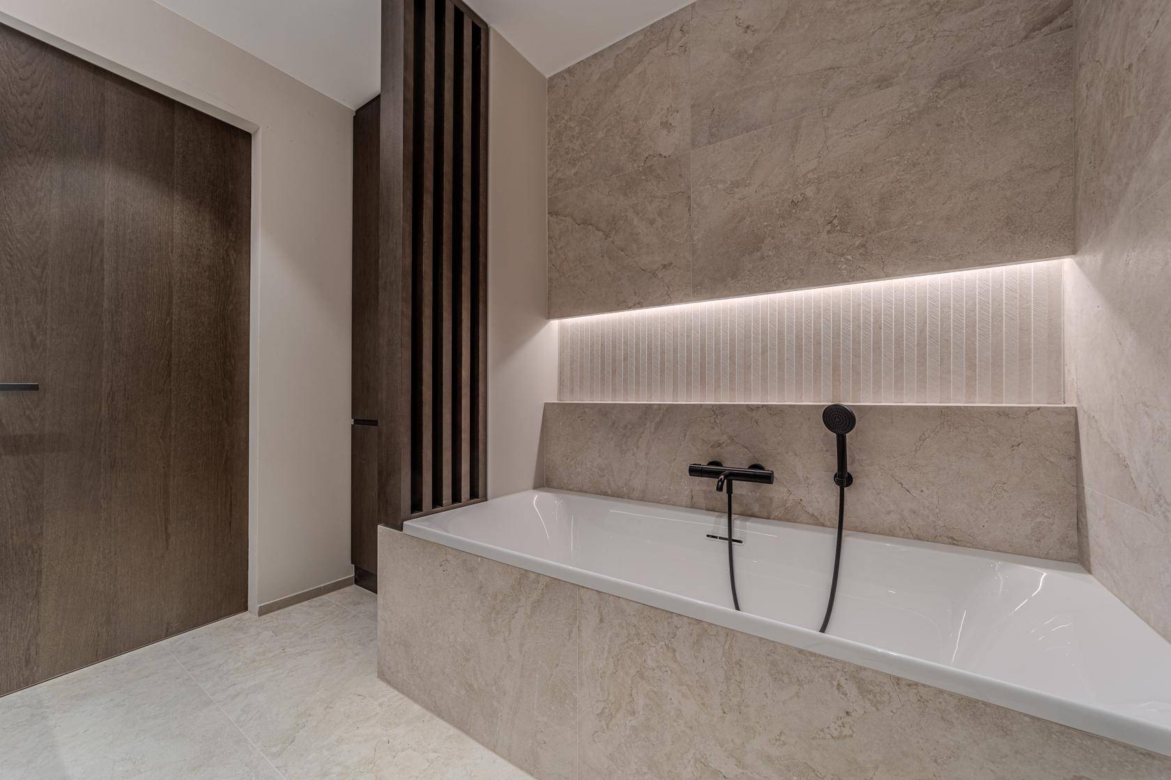 Modernes Badezimmer mit eingebauter Badewanne, beigefarbenen Fliesen und indirekter Beleuchtung.