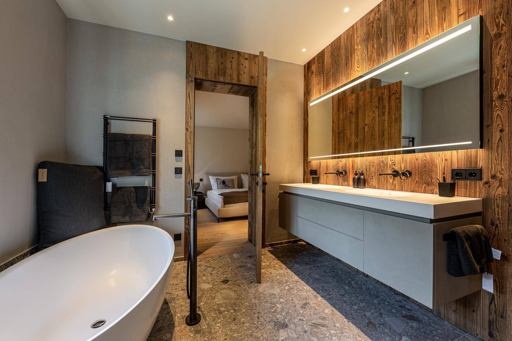 Stilvolles Badezimmer mit Doppelwaschbecken, beleuchtetem Spiegel und freistehender Badewanne, mit Zugang zum Schlafzimmer.