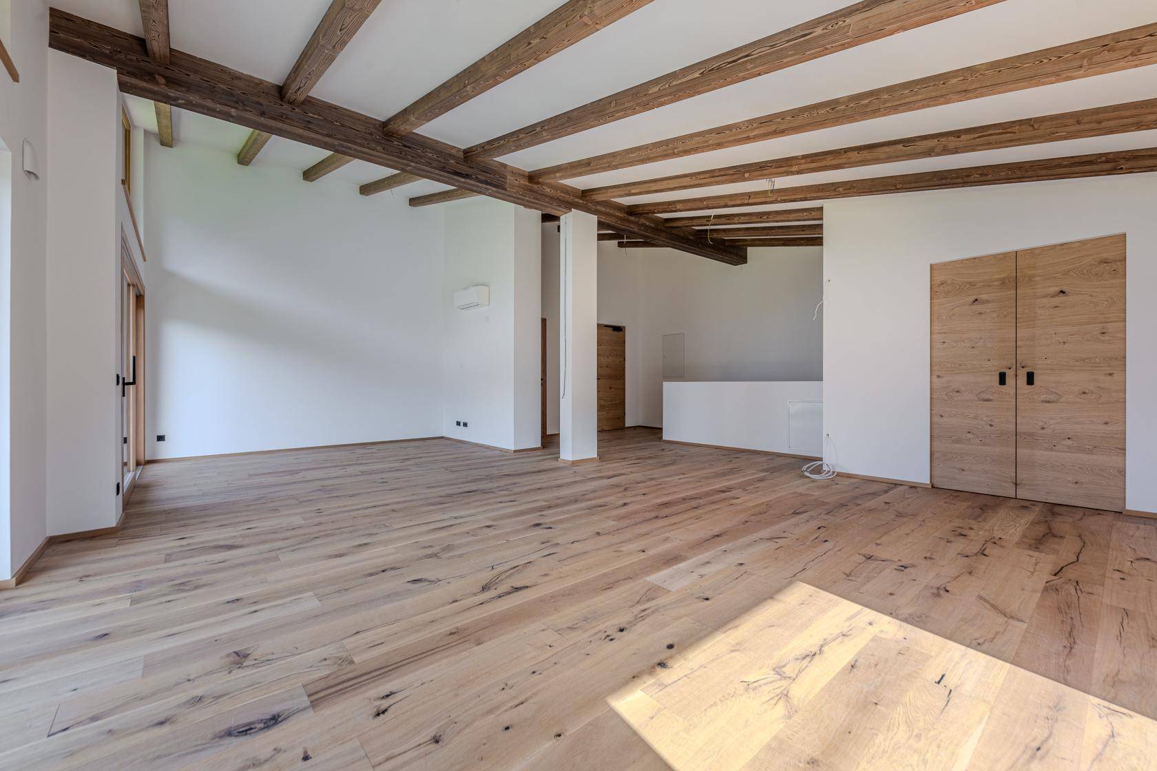 Großer, offener Raum mit Holzboden, sichtbaren Deckenbalken und modernen Holztüren.