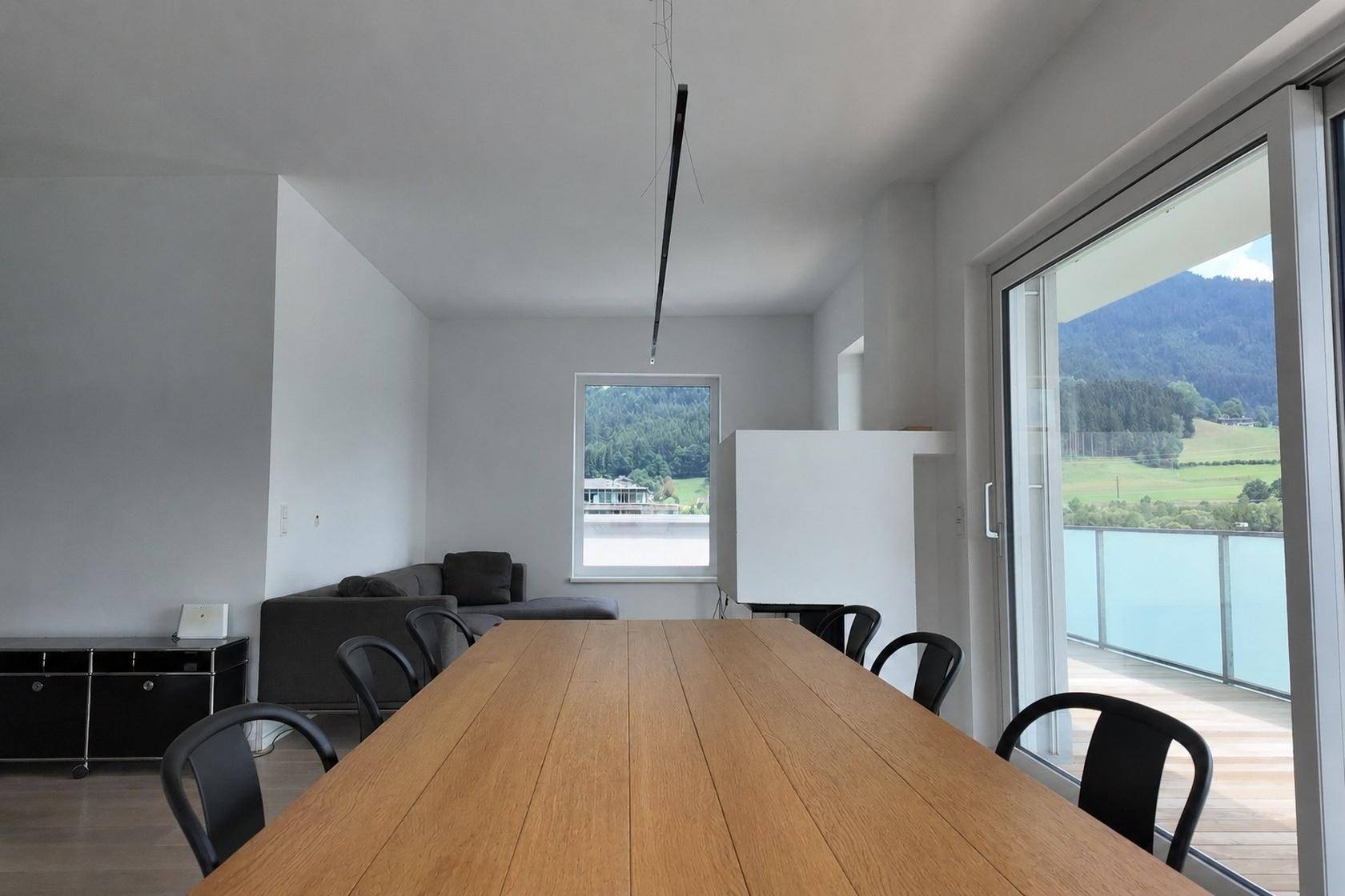 Geräumiger Wohn-Essbereich mit Holztisch, Sofa und großen Fenstern mit Blick auf Berge und See.