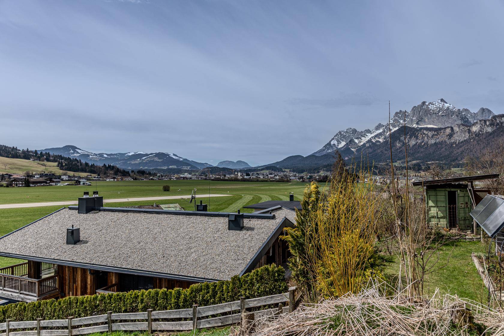 Blick über ein Hausdach auf weite grüne Felder und eine beeindruckende Bergkette in der Ferne.