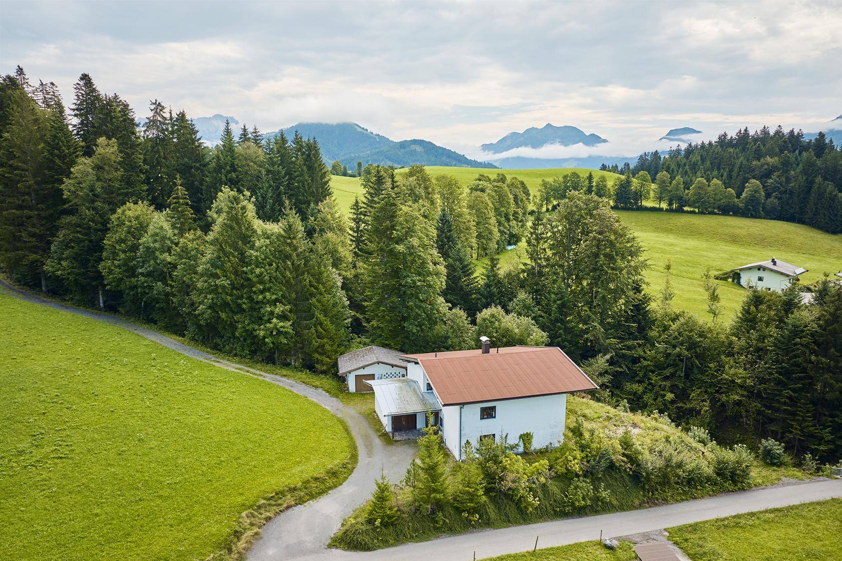 Traditionelles Haus in ländlicher Umgebung mit grünen Wiesen und bewaldeten Bergen im Hintergrund.