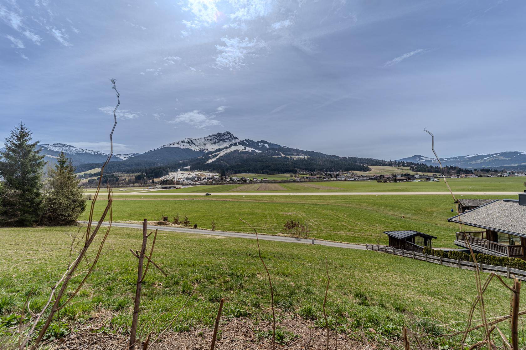 Panoramablick über grüne Felder und eine Straße, mit schneebedeckten Bergen am Horizont unter bewölktem Himmel.