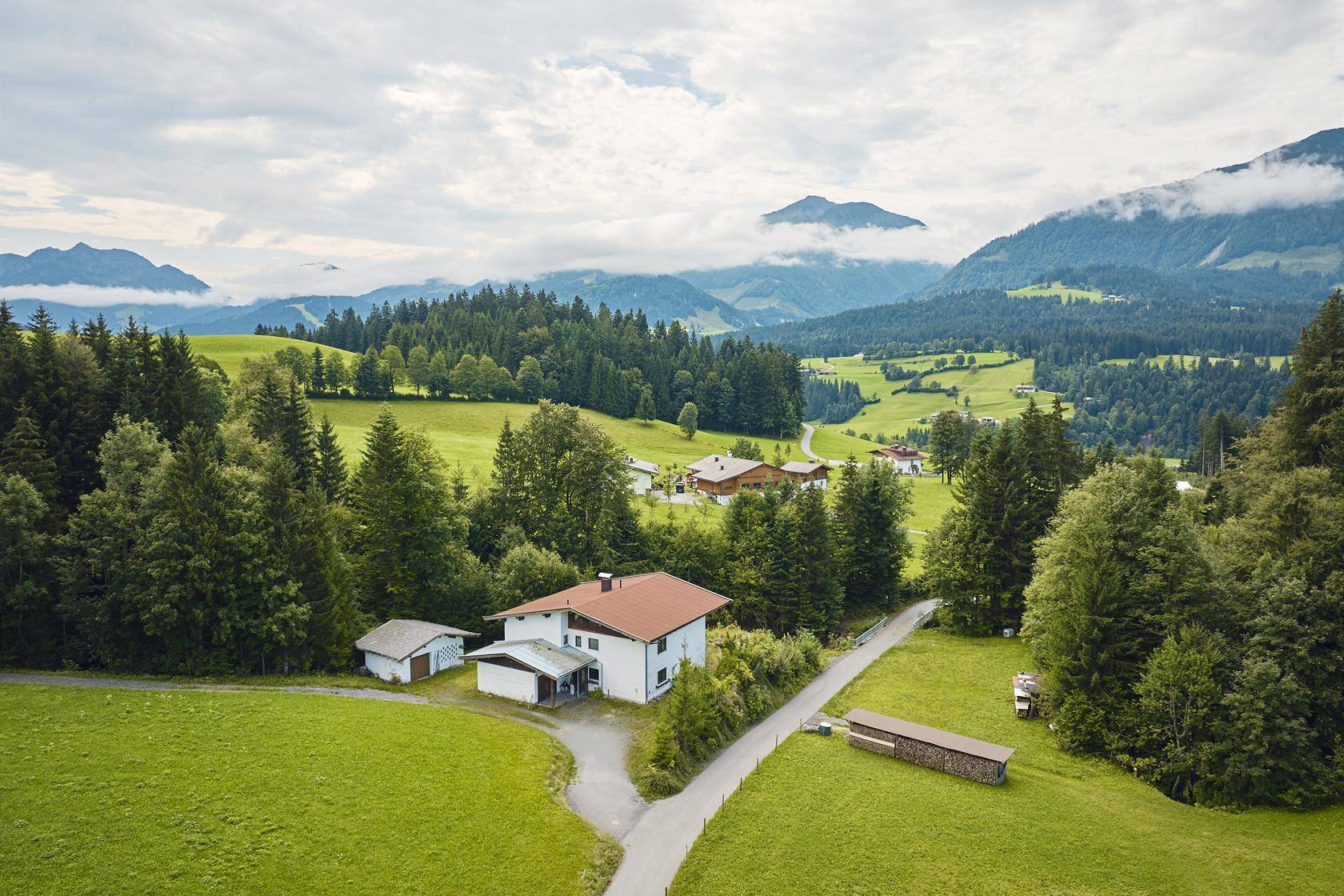 Luftaufnahme eines Hauses in einer ländlichen Siedlung, umgeben von grünen Wiesen und Wäldern.