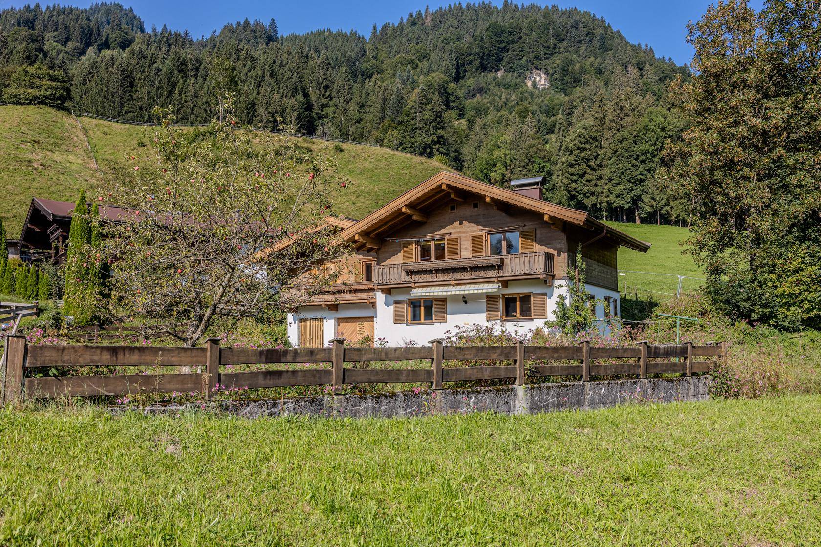 Charmantes Landhaus mit Holzbalkonen und umzäuntem Gartenbereich vor bewaldetem Hang.