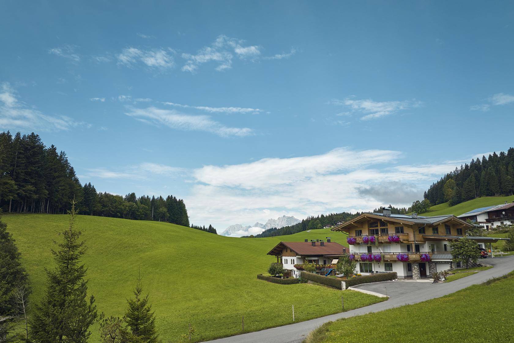 Haus in einer weiten grünen Landschaft mit Bergen im Hintergrund unter blauem Himmel.
