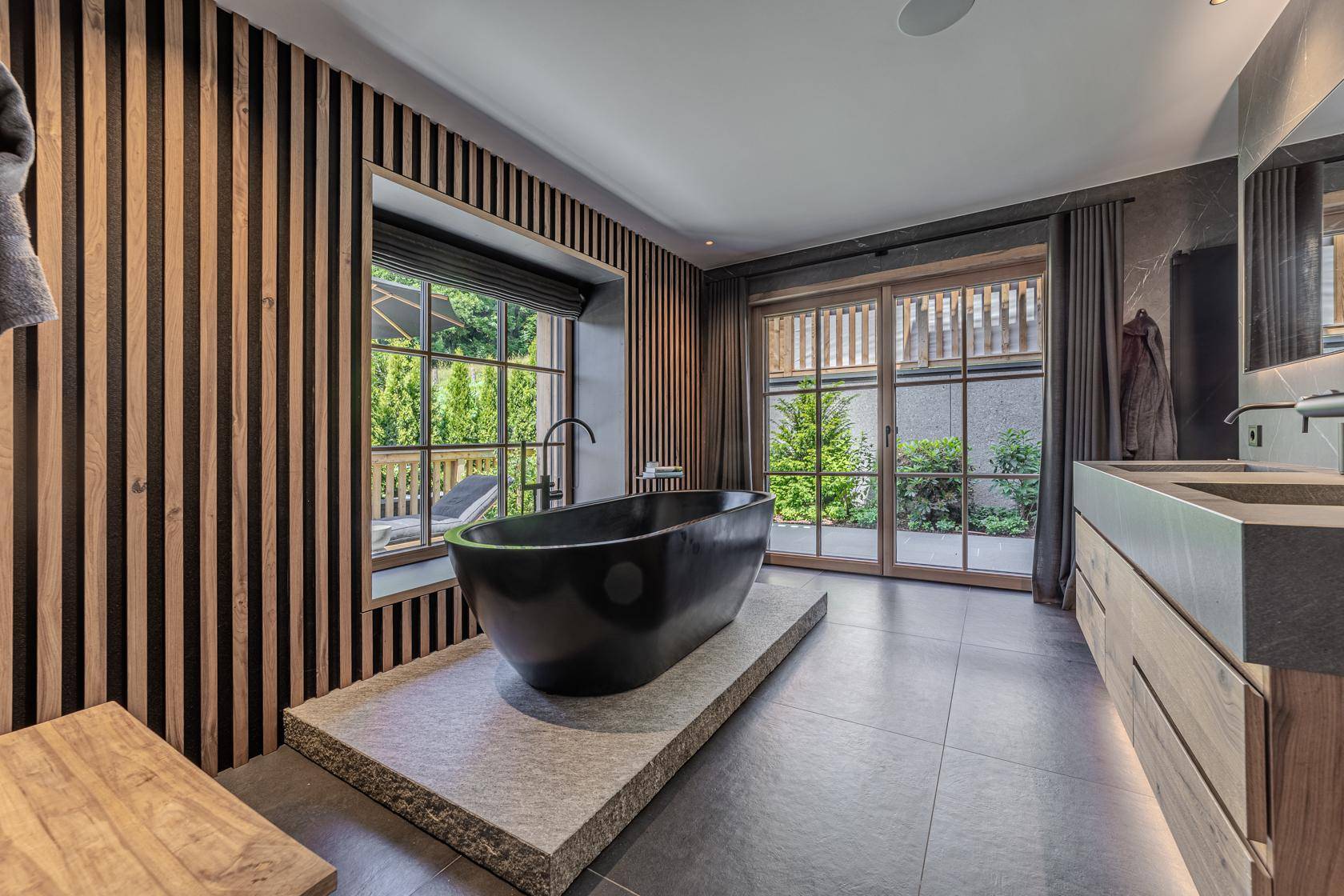 Luxuriöses Badezimmer mit freistehender schwarzer Badewanne, Holzlamellenwand und Zugang zur Terrasse.