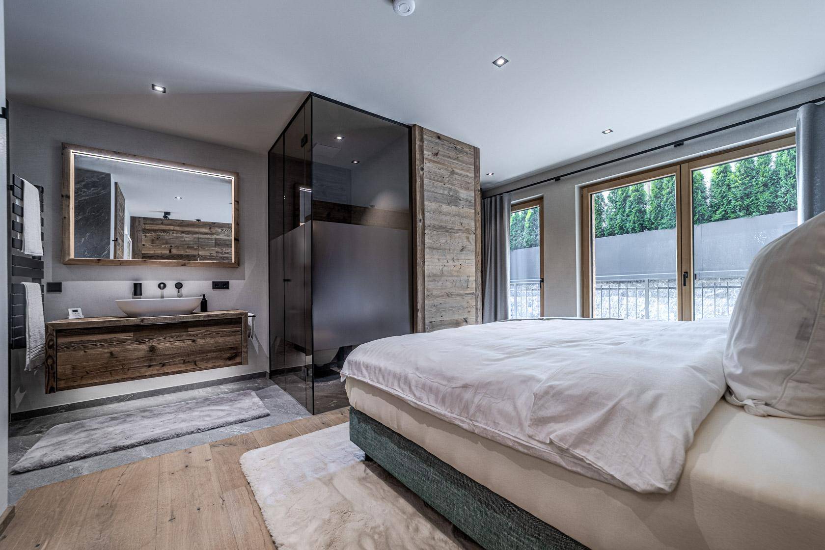 Schlafzimmer mit integriertem Badbereich, Holzboden und großen Fenstern mit Blick ins Grüne.