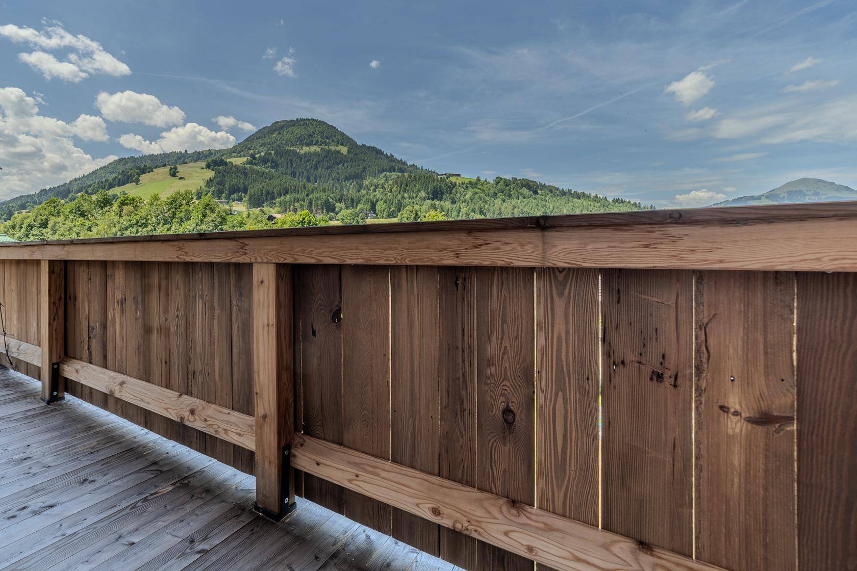 Blick von der Holzterrasse auf die umliegende Berglandschaft unter blauem Himmel.