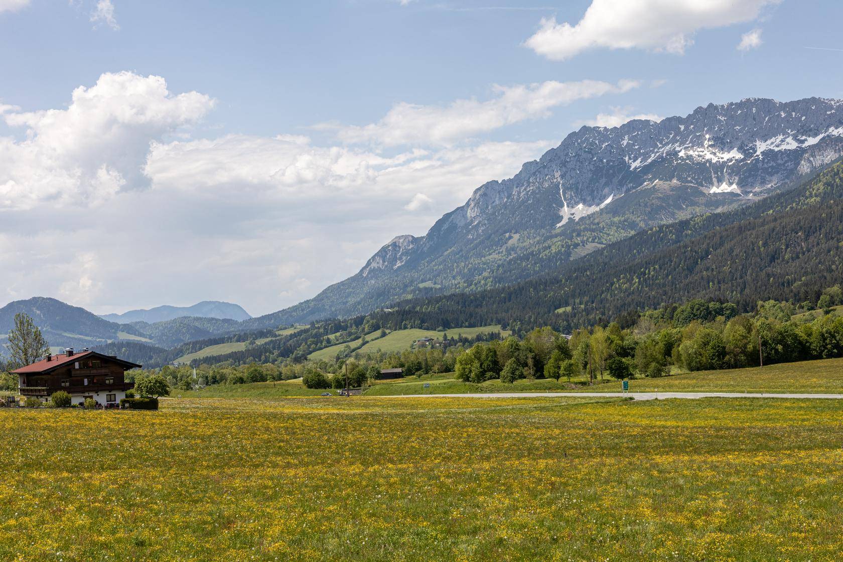 Weitläufige ländliche Landschaft mit grünen Wiesen, einem traditionellen Haus und majestätischen Bergen.