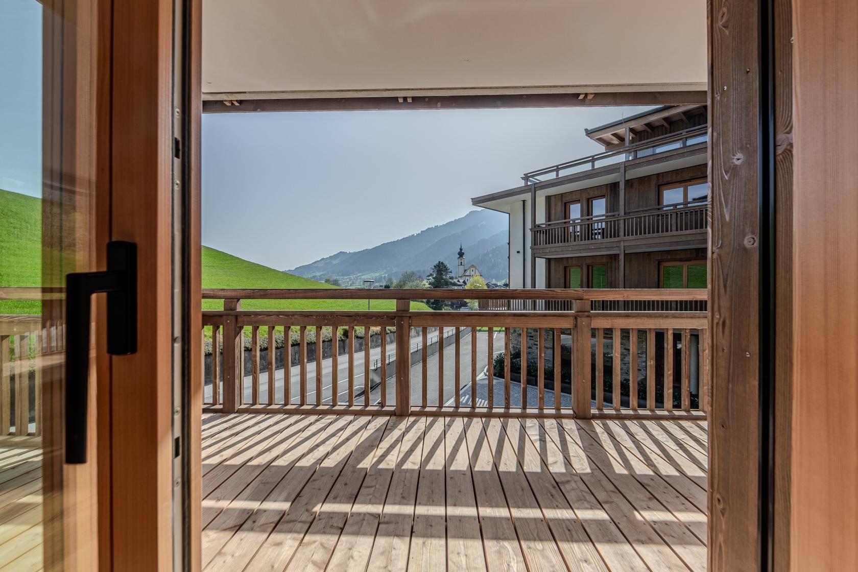 Holzbalkon mit traditionellem Geländer und sonnigem Ausblick auf die umliegende Berglandschaft und Nachbargebäude.