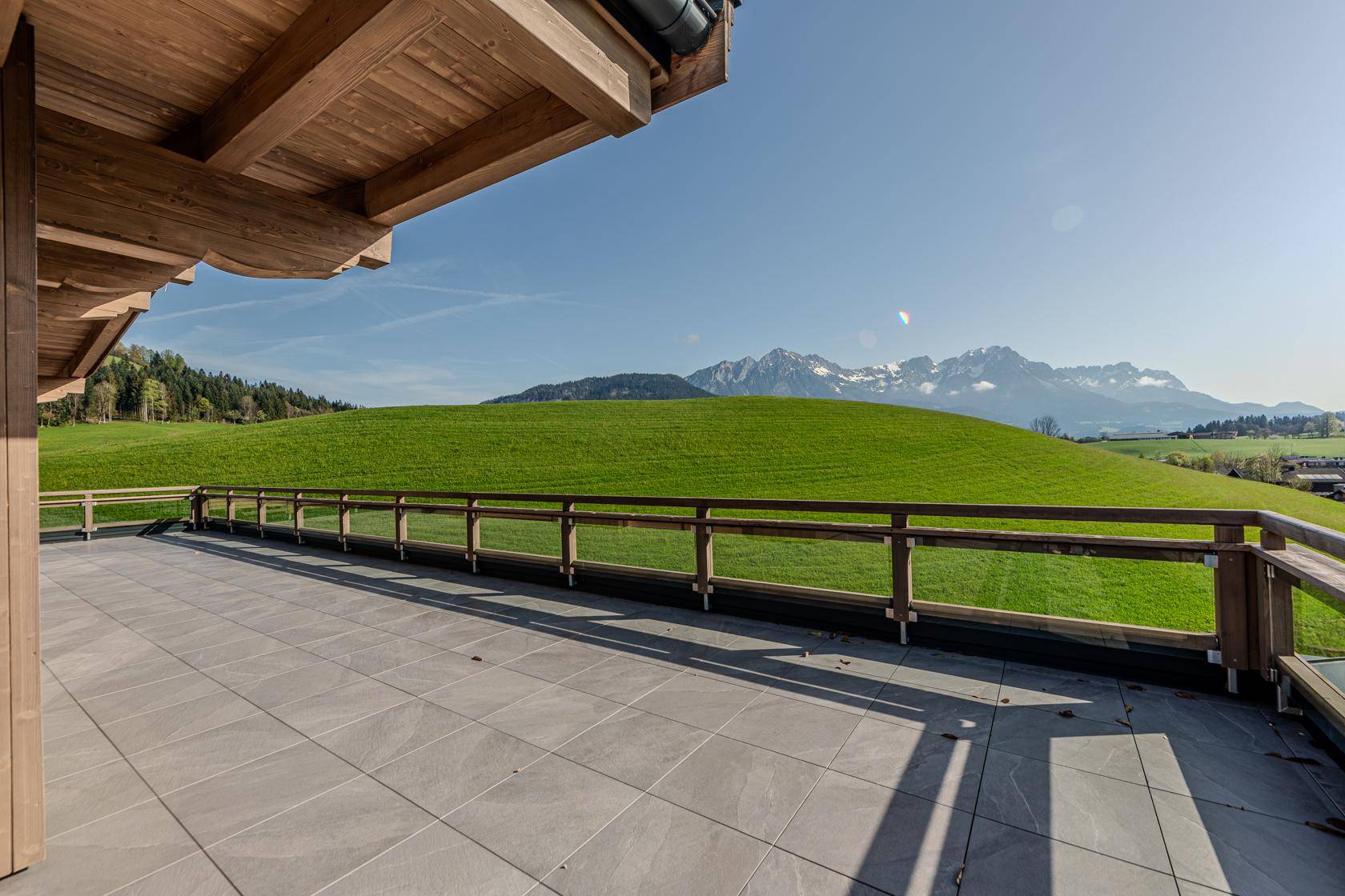 Große geflieste Terrasse mit Holzgeländer und weitem Blick auf grüne Hügel und Berge.