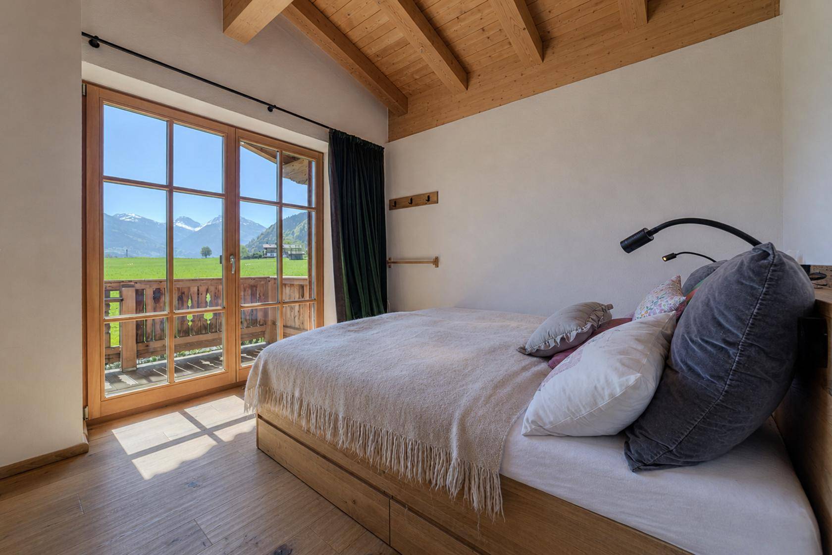 Helles Schlafzimmer mit Holzbalkendecke, Balkonzugang und Blick auf die Berge und Wiesen.