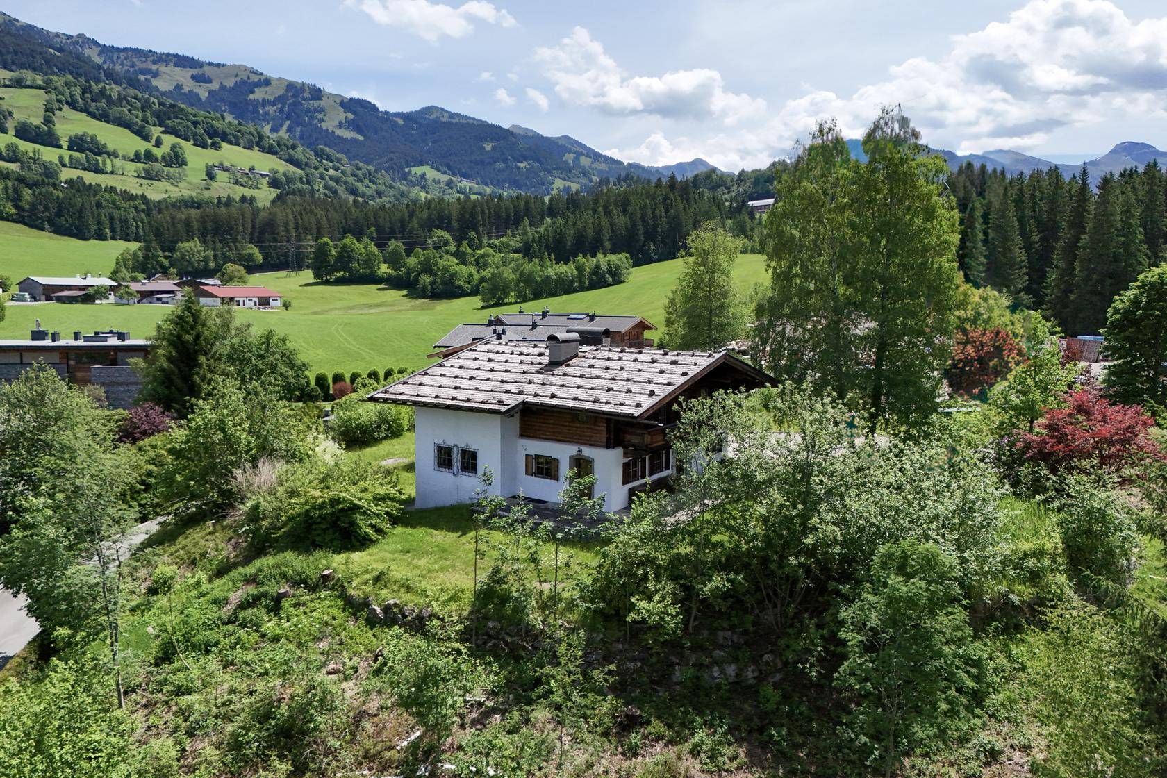 Charmantes Haus in alpiner Landschaft, umgeben von grünen Hügeln und Bergen unter blauem Himmel.