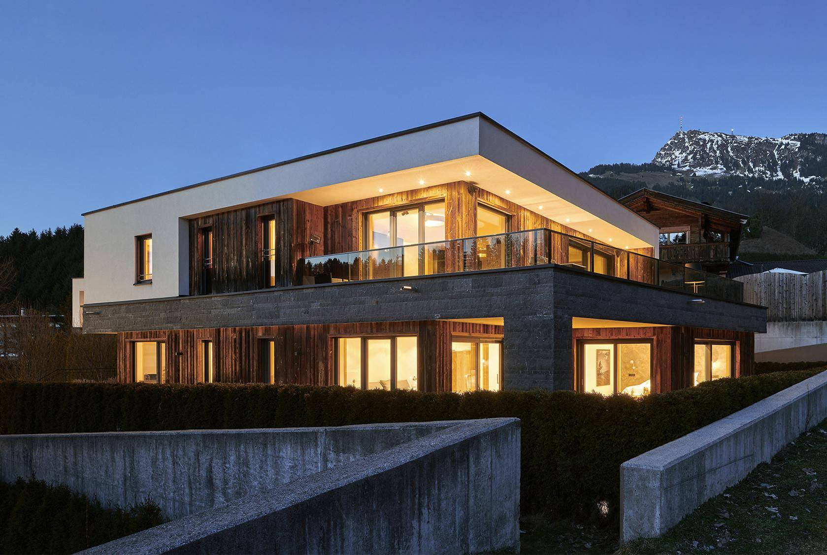 Moderne Architektur mit Holzfassade, großen Fenstern und Balkonen vor alpiner Bergkulisse.