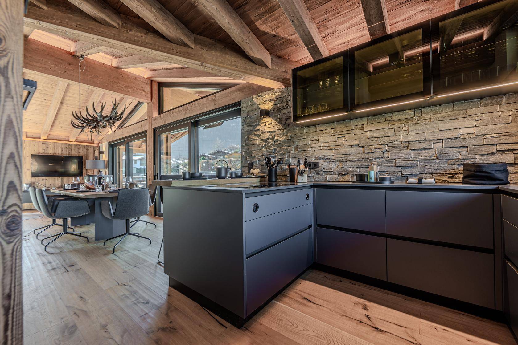 Moderne, offene Küche mit dunklen Schränken, Steinwand und Holzbalkendecke, integriert in den Wohnbereich.