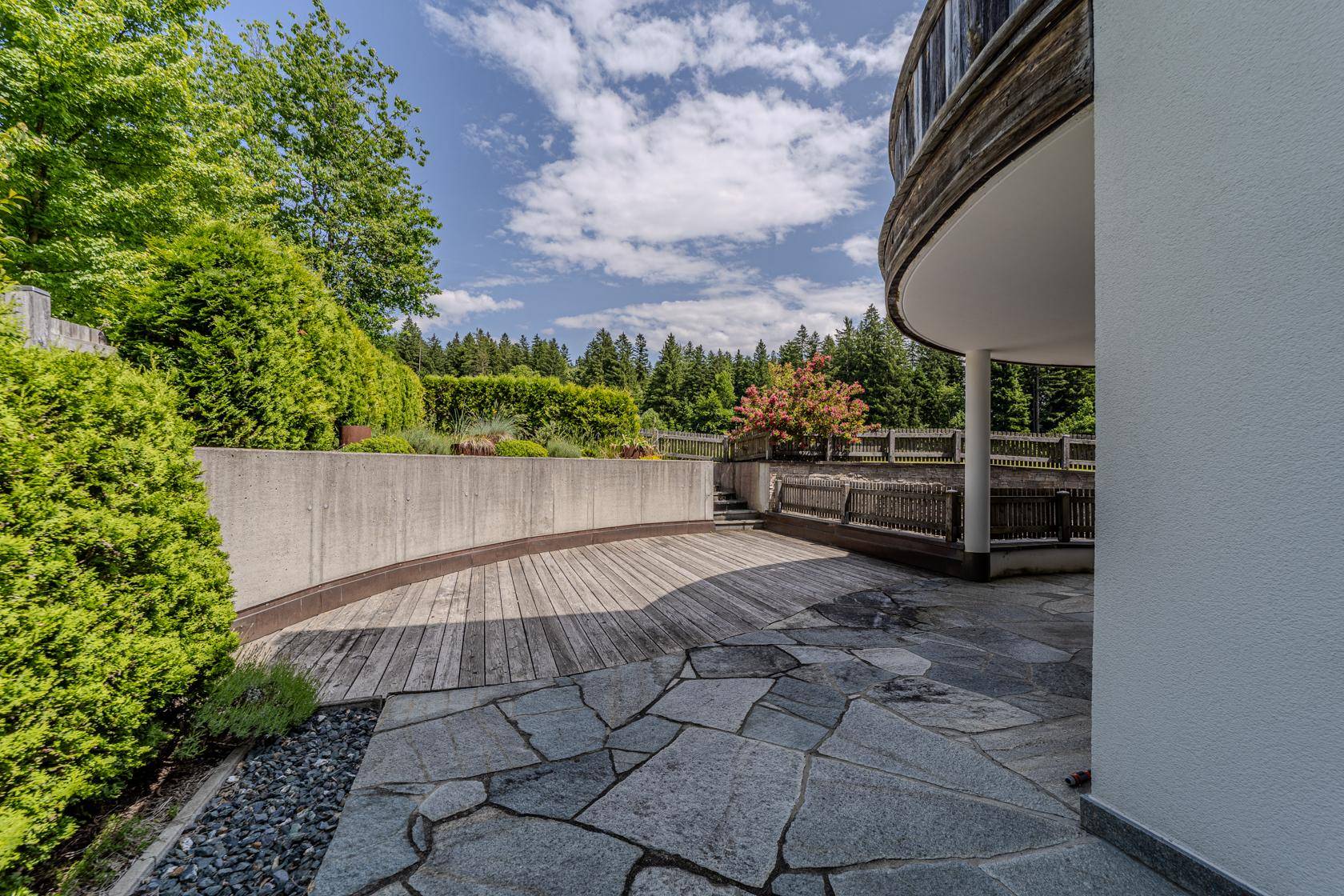 Große Steinterrasse mit Holzdeck, umgeben von Grün und Bäumen.