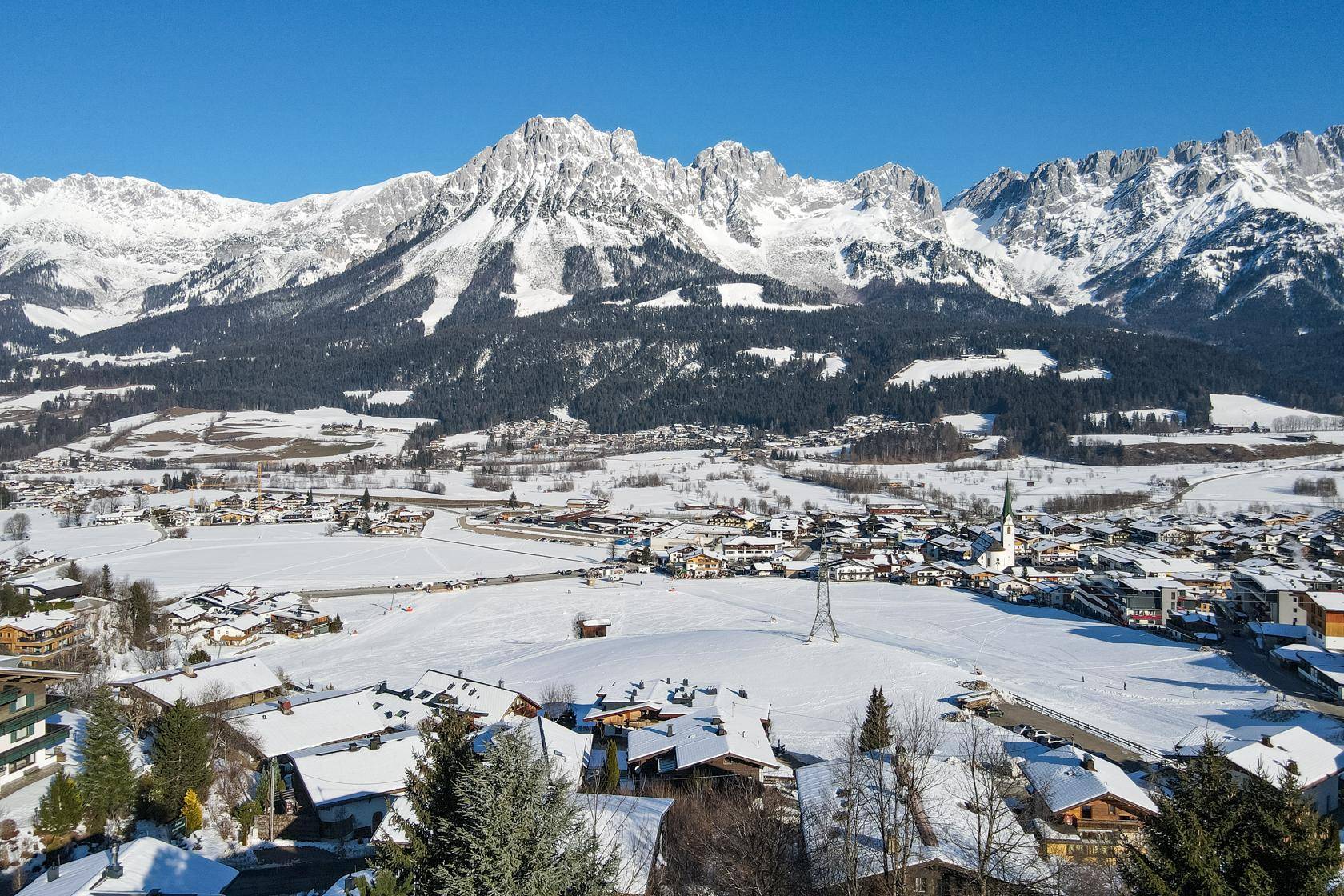 Winterlandschaft mit schneebedeckten Bergen und einem verschneiten Dorf unter strahlend blauem Himmel.