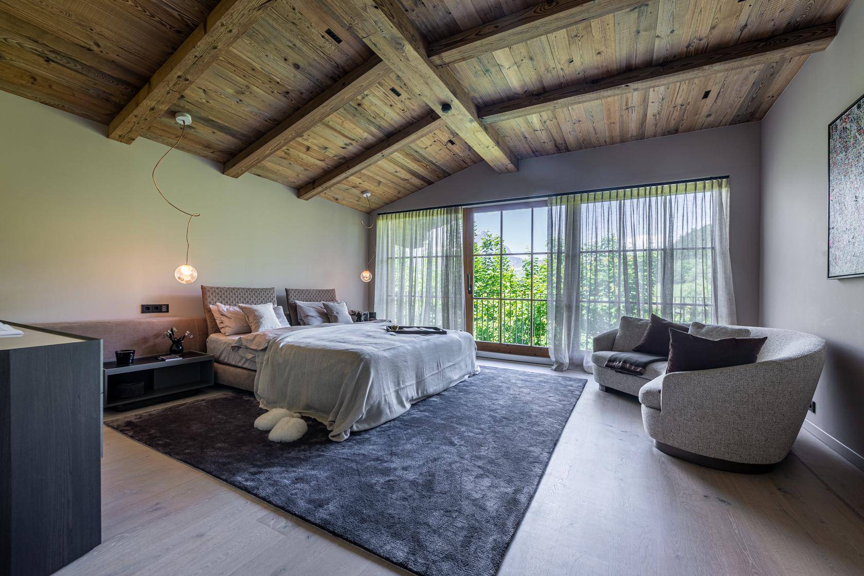 Helles Schlafzimmer mit hoher Holzdecke, freiliegenden Balken und großen Fenstern mit Blick auf die Natur.