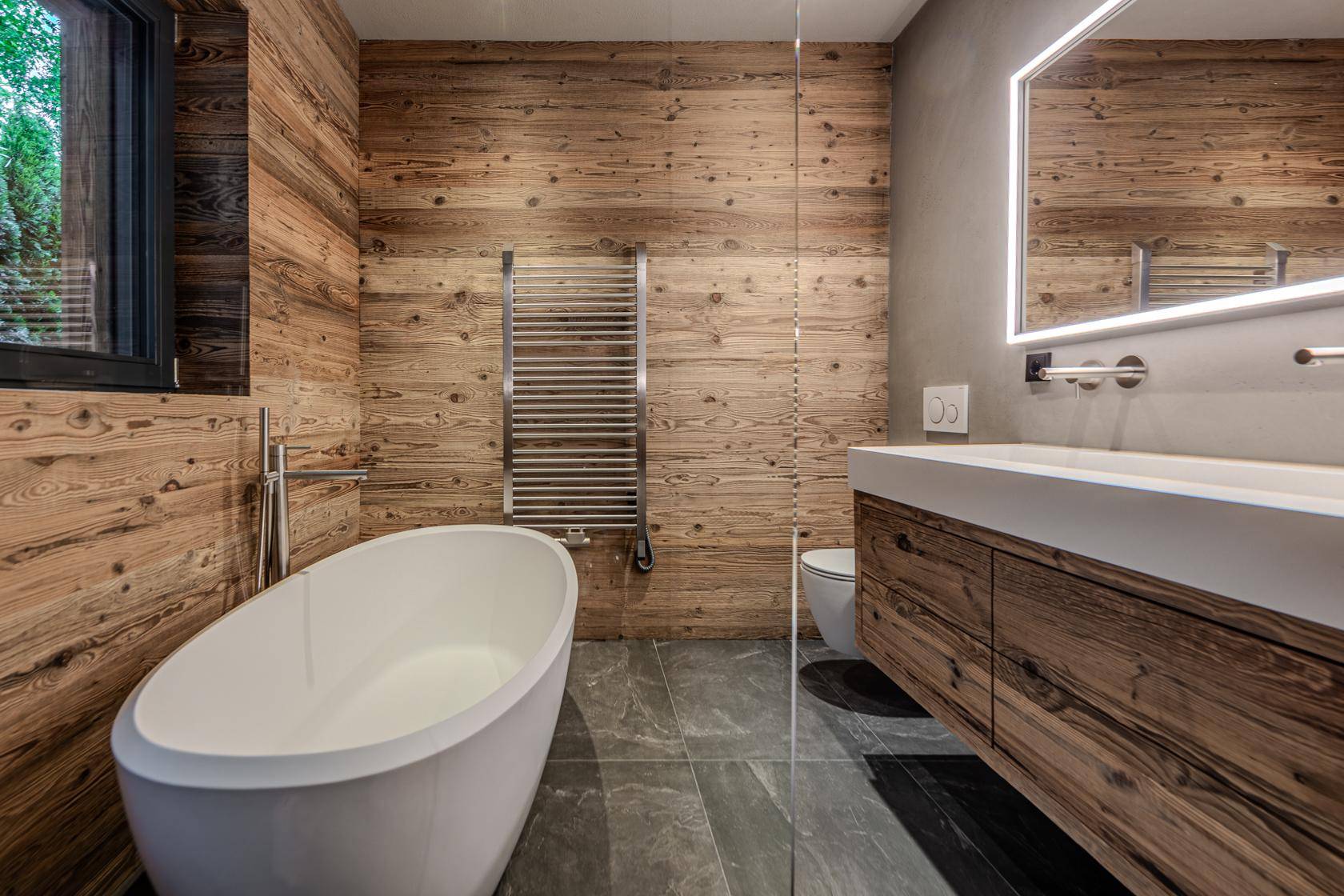 Modernes Badezimmer mit freistehender Badewanne, Holzverkleidung und beleuchtetem Spiegel.