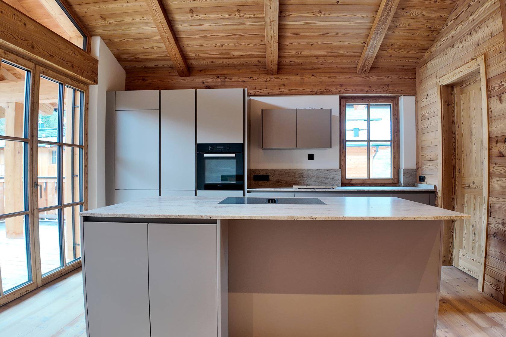 Moderne Einbauküche mit Kochinsel, integrierten Geräten und Zugang zu einem Balkon durch große Fenster.
