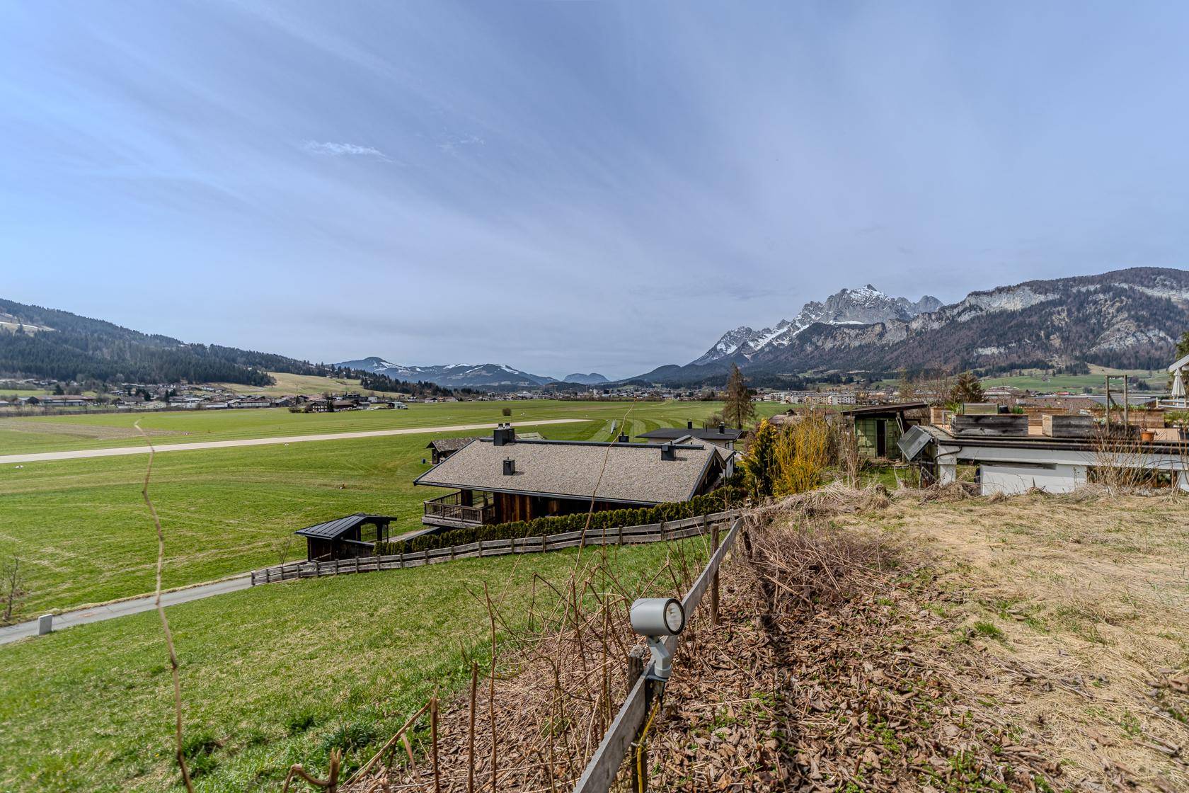 Weitläufige grüne Landschaft mit einem Haus im Vordergrund und schneebedeckten Bergen am Horizont.